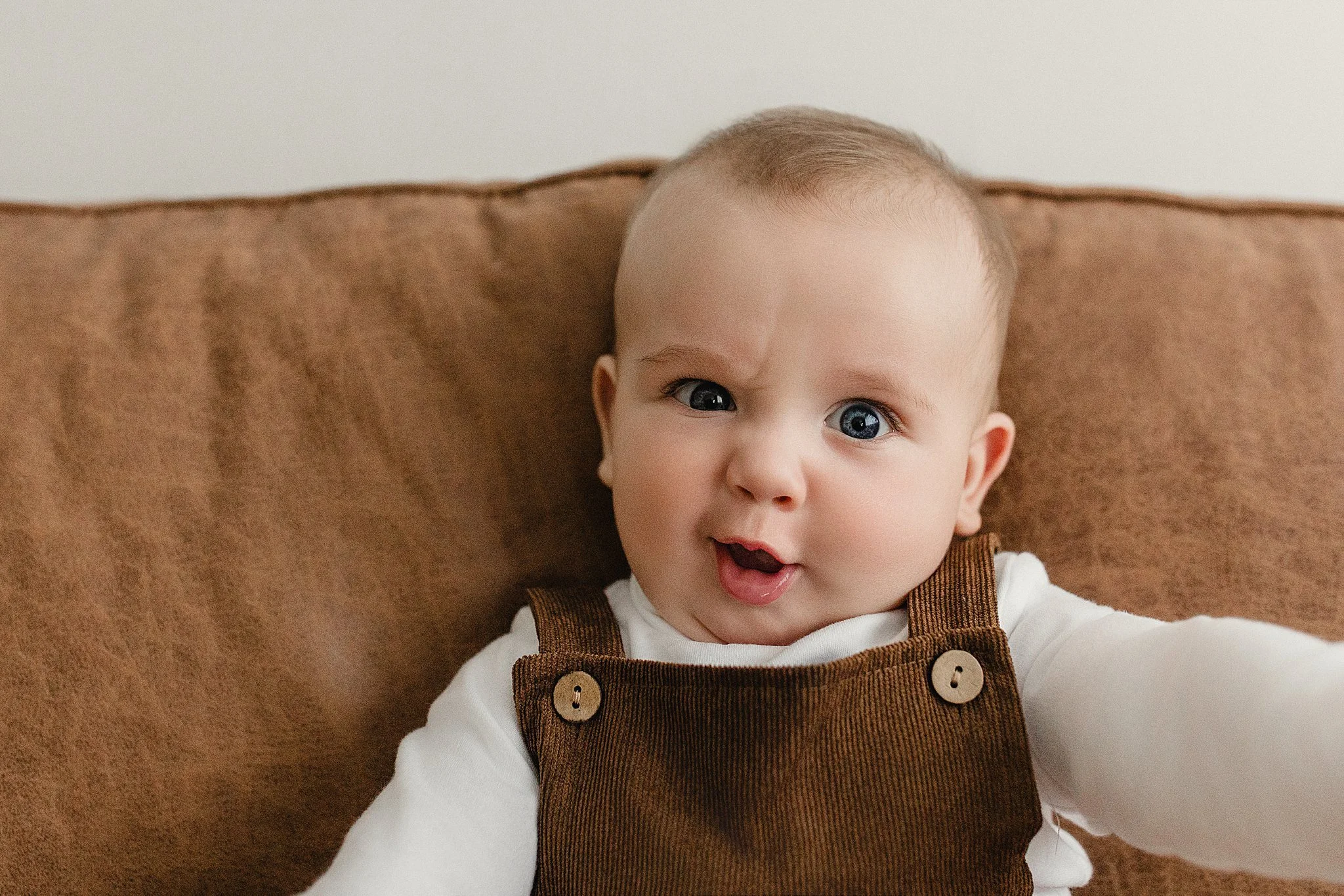 A baby with blue eyes and light hair, wearing a white shirt and brown overalls, sitting on a brown couch, making a surprised facial expression during lifestyle baby photos in Naperville.