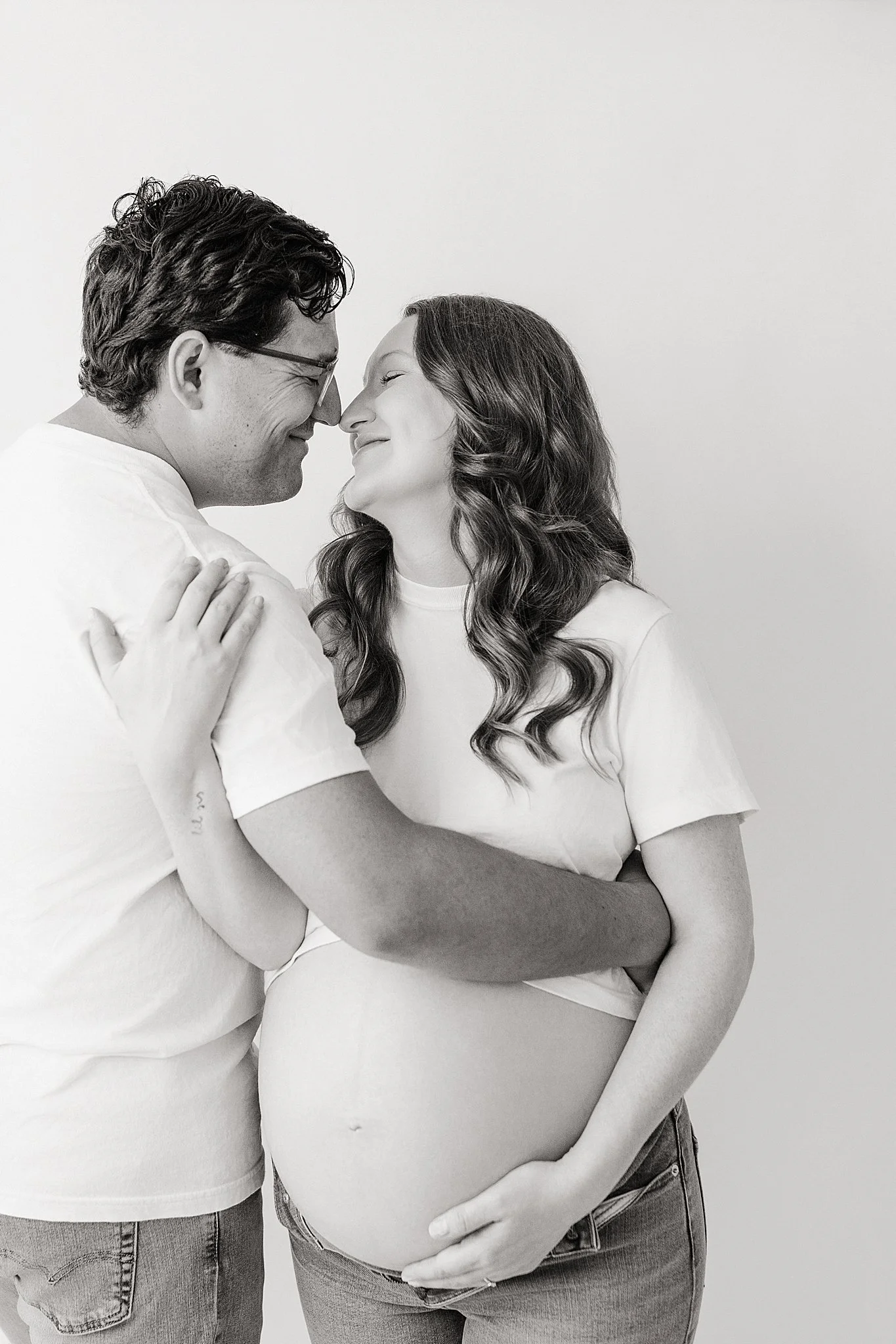 A black and white photo of a man and pregnant woman in a close embrace, touching foreheads and smiling, with the man's hand resting on her arm and her hand on her belly during maternity photos near me.