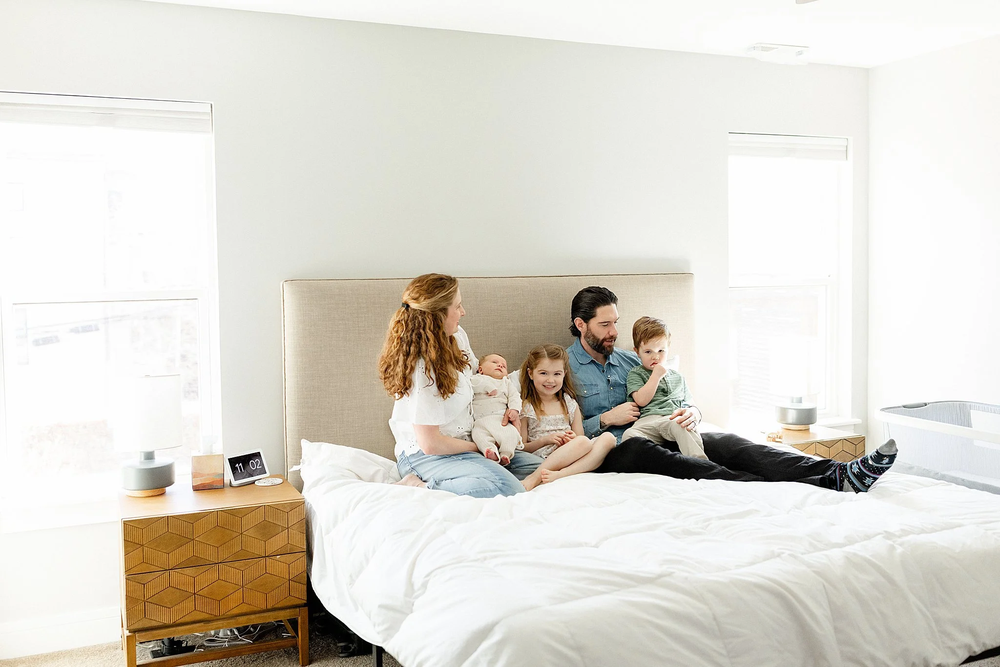 Family sitting on bed in a bright, modern bedroom with large windows, white walls, and minimal decor with lifestyle photographer Ally and B Photography.