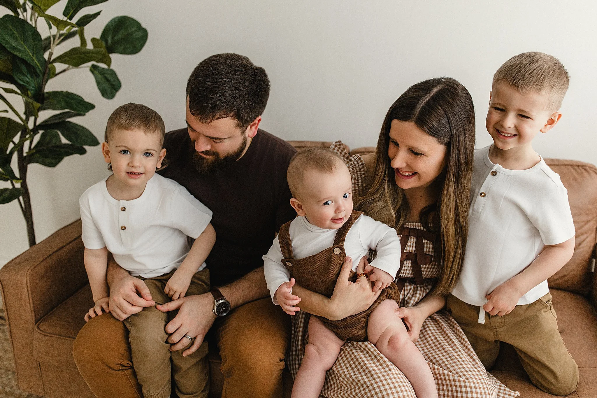 Family of five sitting on a brown couch in a living room, smiling and enjoying each other's company during lifestyle photos with Ally and B Photography in Naperville, IL.