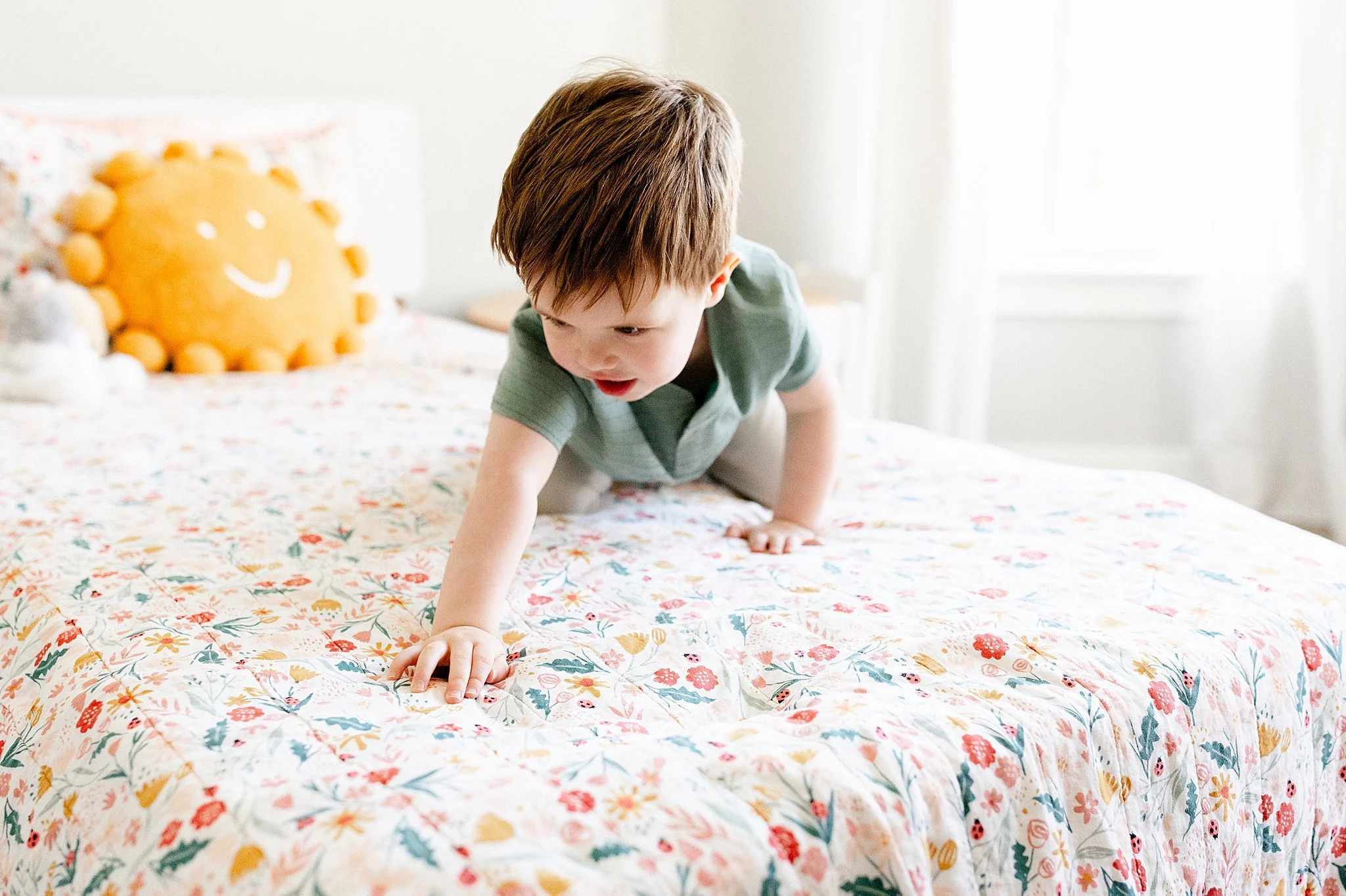 A young boy crawling on a bed with a floral bedspread, near a decorative cushion with a smiling face, in a bright room with white walls and a window.
