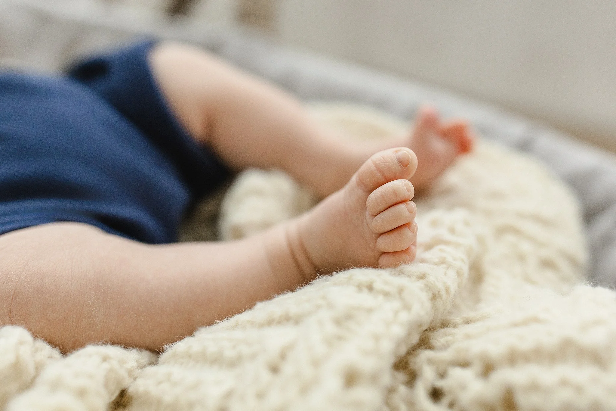 Close-up of a baby lying on a soft cream blanket, showing the baby's hand and partially visible arm, wearing a dark blue outfit during a baby photoshoot near me.