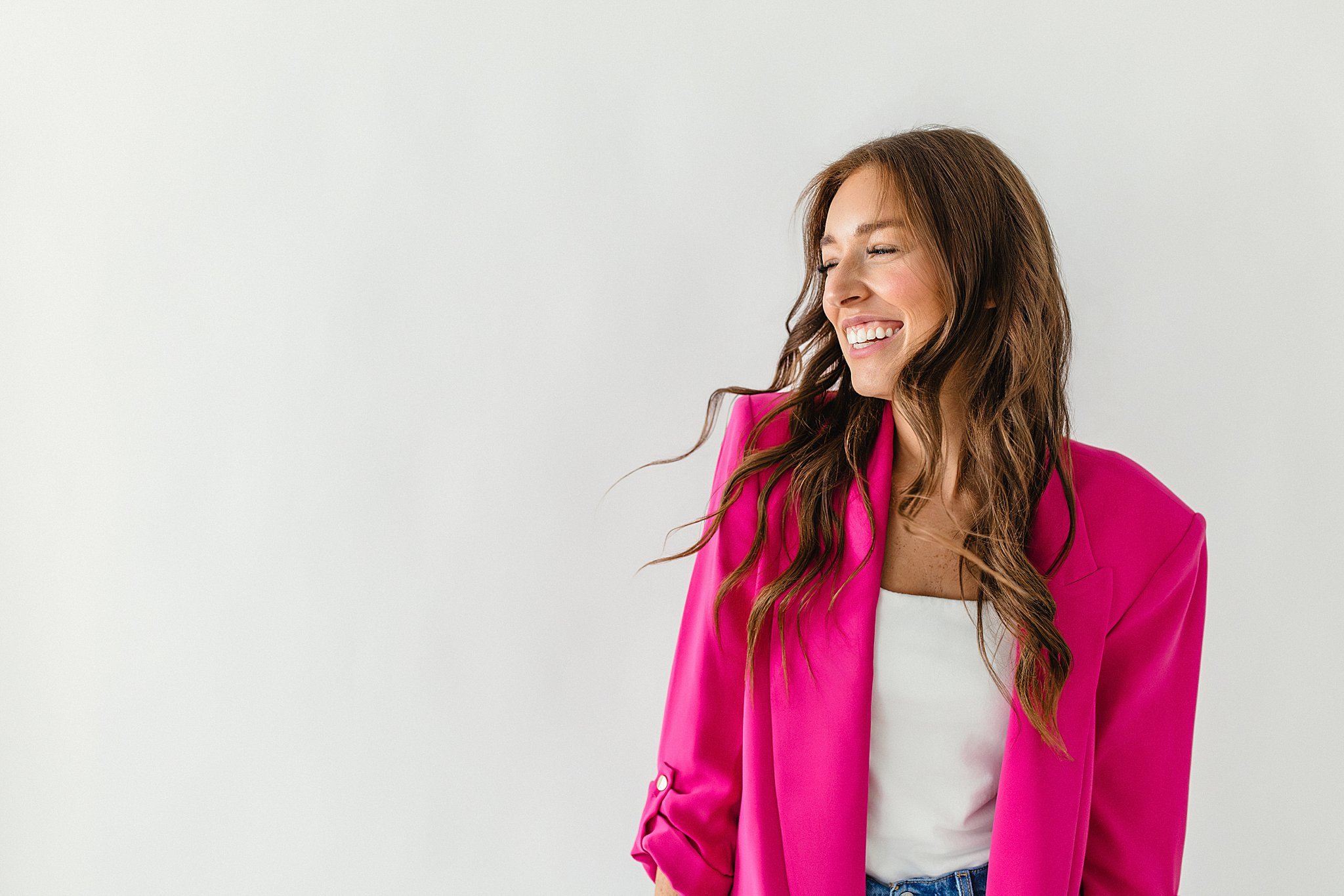 A woman with long wavy brown hair wearing a bright pink blazer and a white top, smiling and looking to her left against a plain white background for personal branding photos in Naperville, IL.