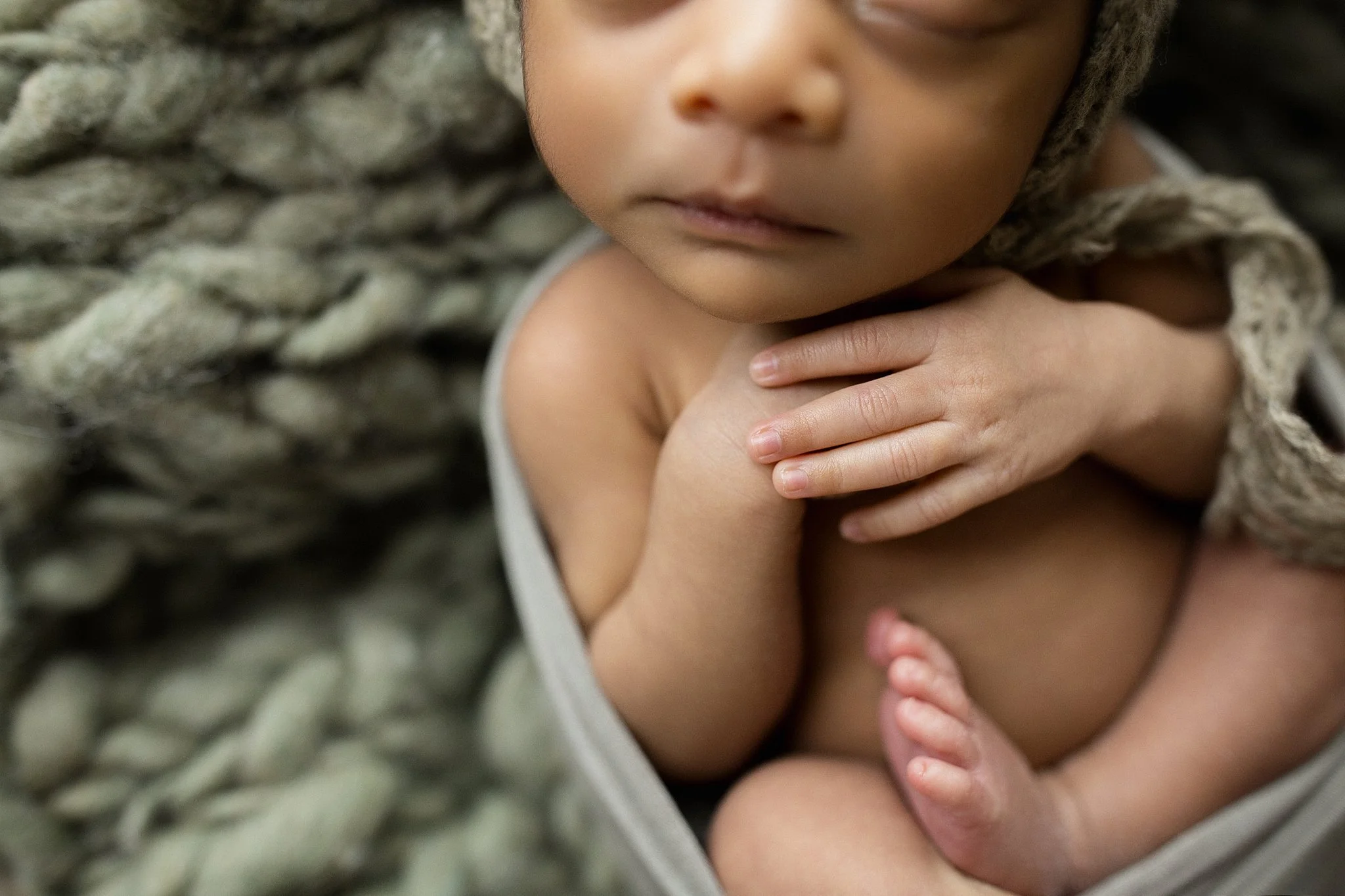 Close-up of a young child with hands resting on their chest, sitting on a textured woven blanket during newborn photos in downtown Naperville, IL.