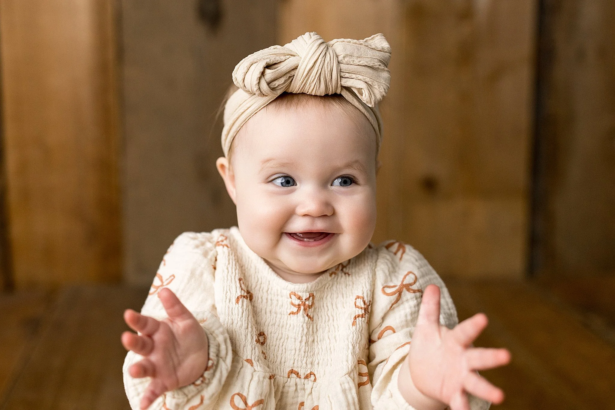 Happy young child with blue eyes, wearing a beige headwrap and a cream-colored sweater with brown patterns, reaching forward with both hands with baby photographers near me.