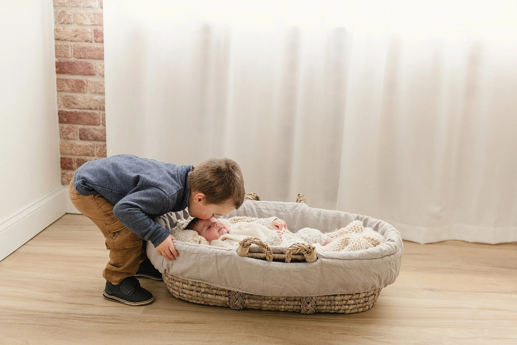 A young boy leaning over and kissing a newborn baby lying in a basket on the floor during lifestyle family photos with Ally and B Photography in Naperville, IL.