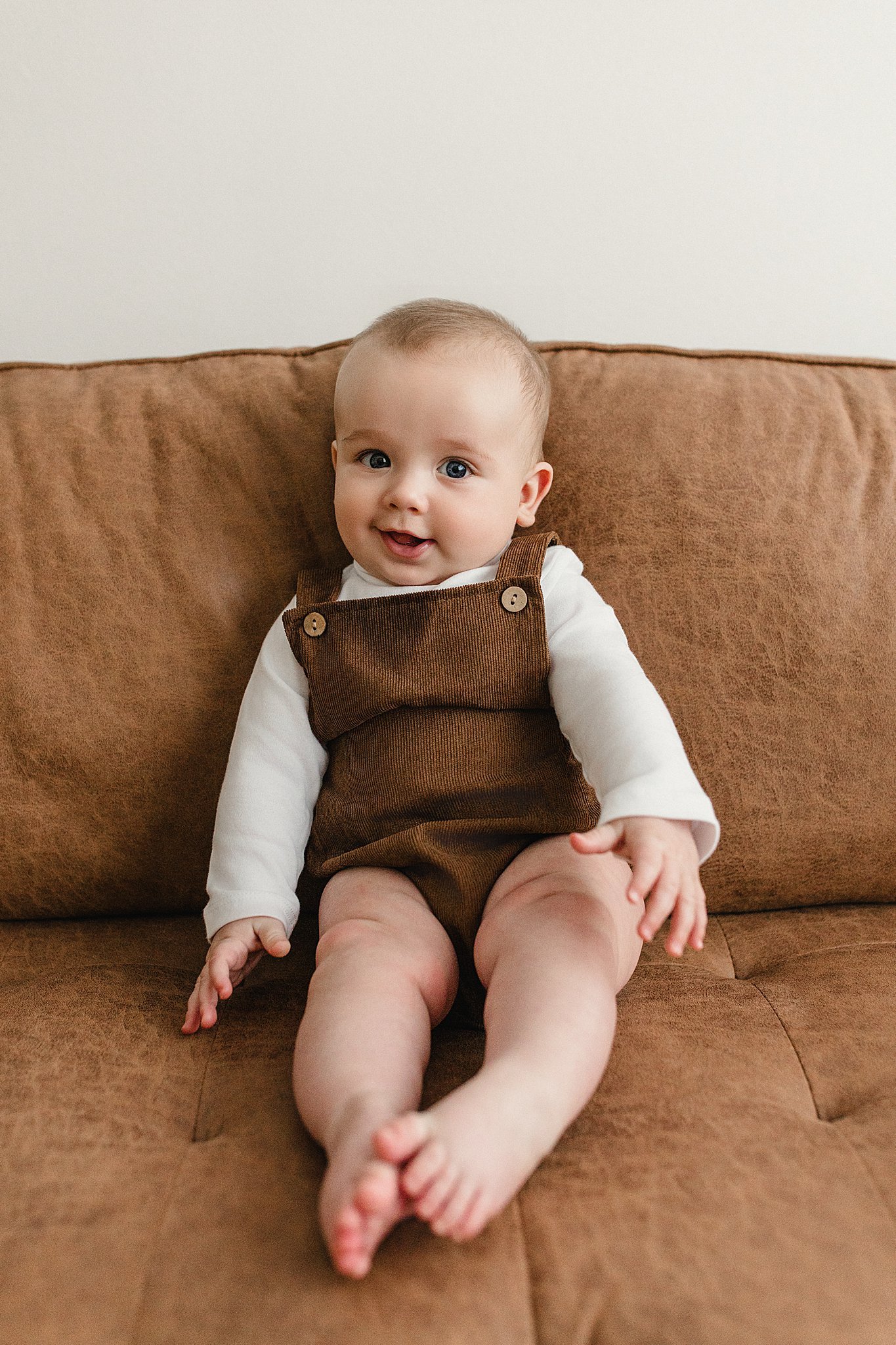 A smiling baby sitting on a brown sofa, wearing a white long-sleeve shirt and brown overalls with best lifestyle family photographer near me.