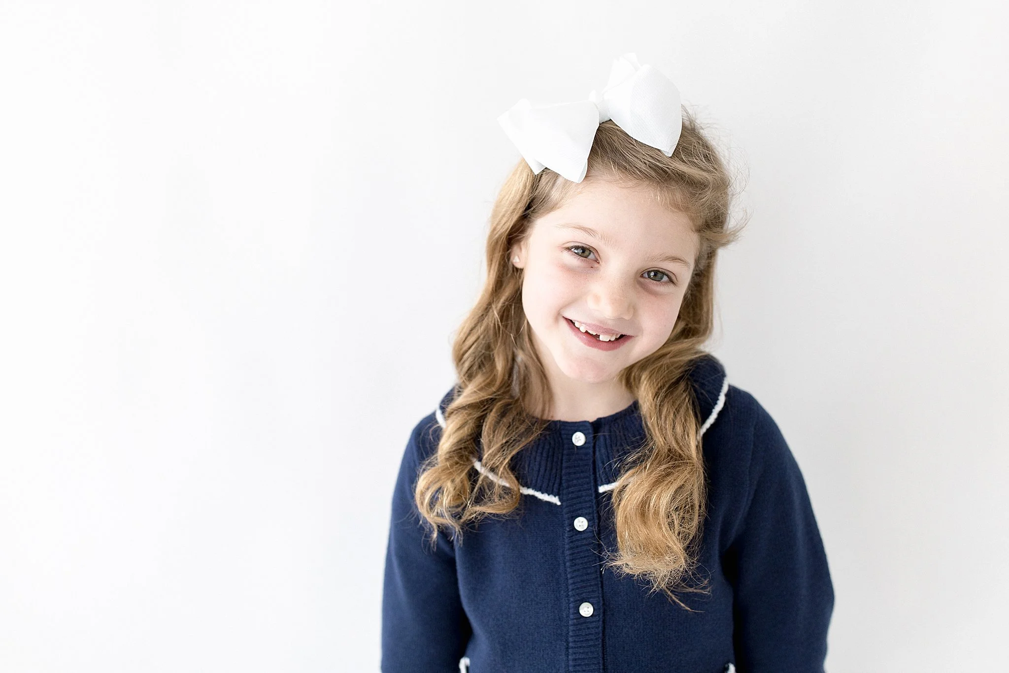 A young girl with long curly blonde hair, wearing a dark blue cardigan with white accents and a large white bow in her hair, smiling against a plain white background during a kids portrait session in naperville, IL.