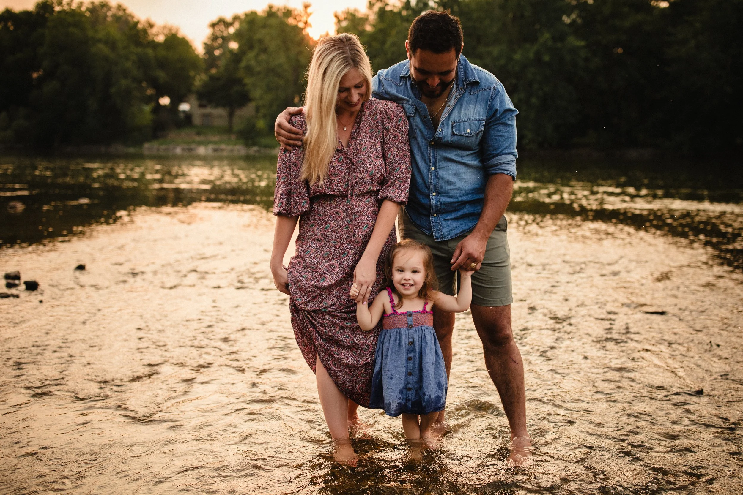 A family of three, including a woman, a man, and a young girl, wading in a river during sunset. They are holding the girl’s hands, and they are smiling and looking down at the water, with trees the sunset during a family creek photoshoot.