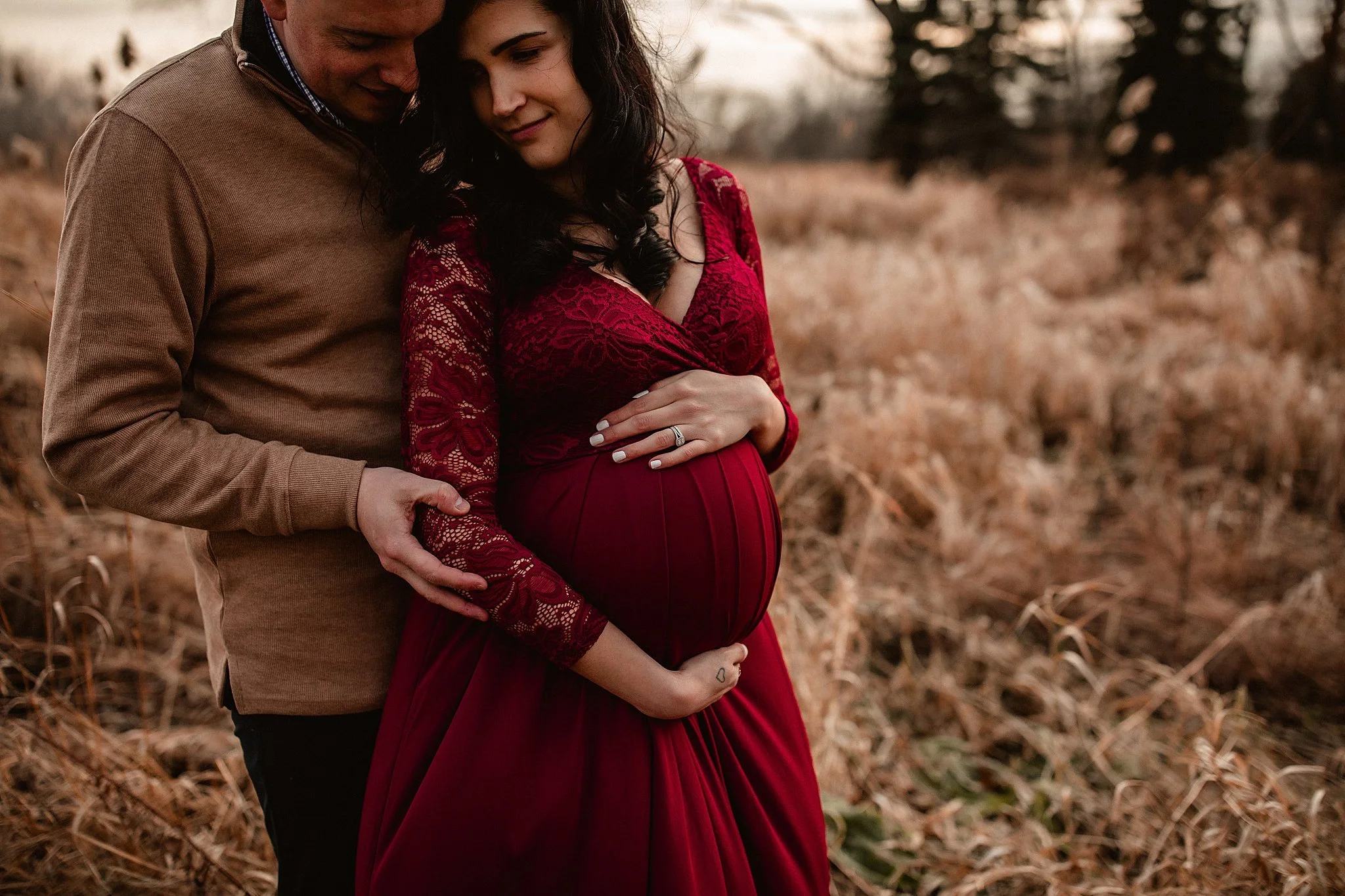 A couple standing in a field, with the woman pregnant and wearing a red lace dress, and the man holding her belly gently during maternity photography Naperville.