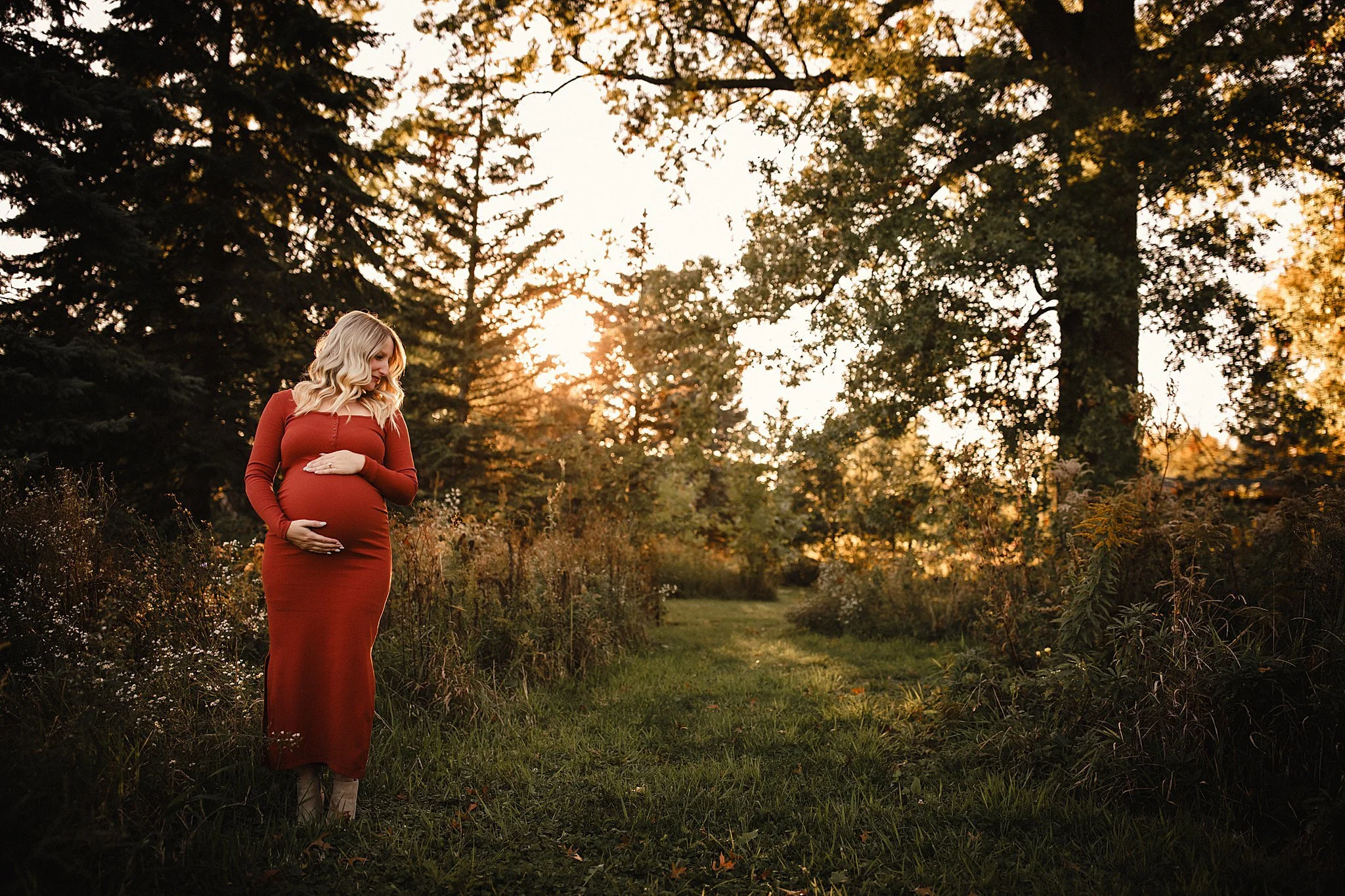 A pregnant woman in a long red dress standing in a grassy path surrounded by trees at sunset with maternity photographers Naperville.