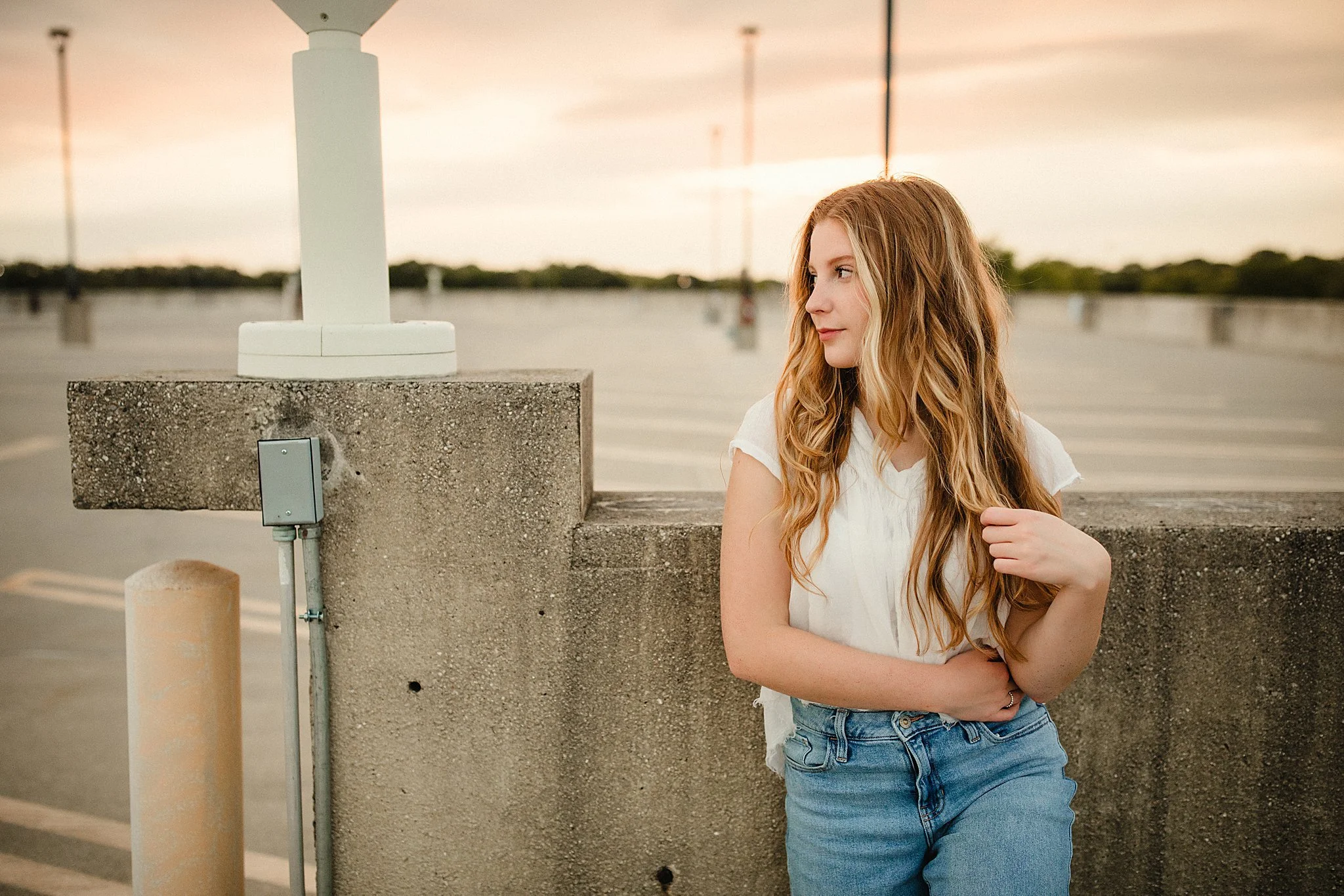 A young woman with long, wavy red hair wearing a white top and blue jeans, standing against a concrete wall in an empty rooftop parking lot at sunset during Naperville senior photos.