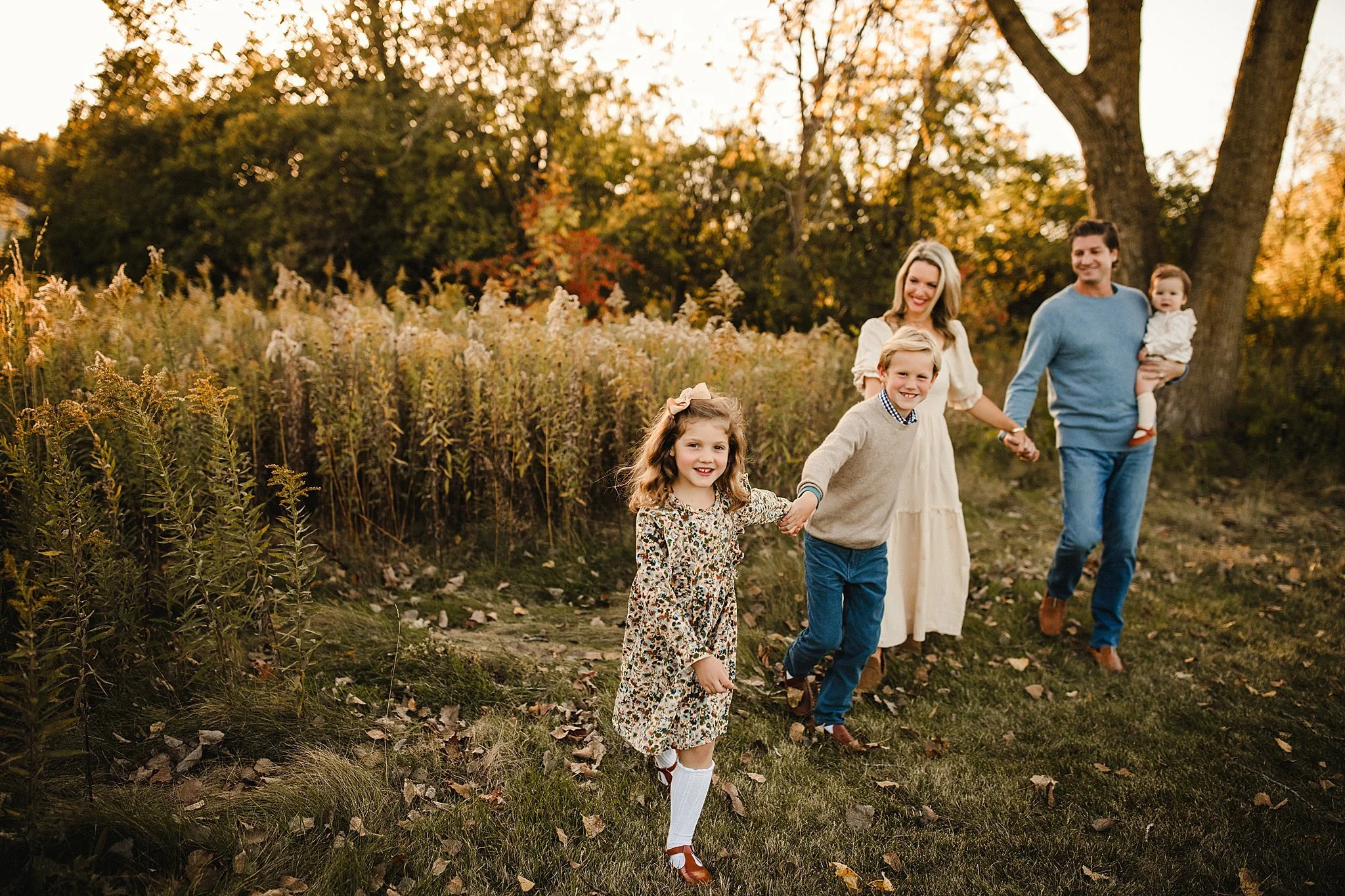 A family of five holding hands and walking outdoors in a park during autumn, with trees and fallen leaves around for a fall family photo session in Naperville with Marie Lopez of Ally and B Photography.