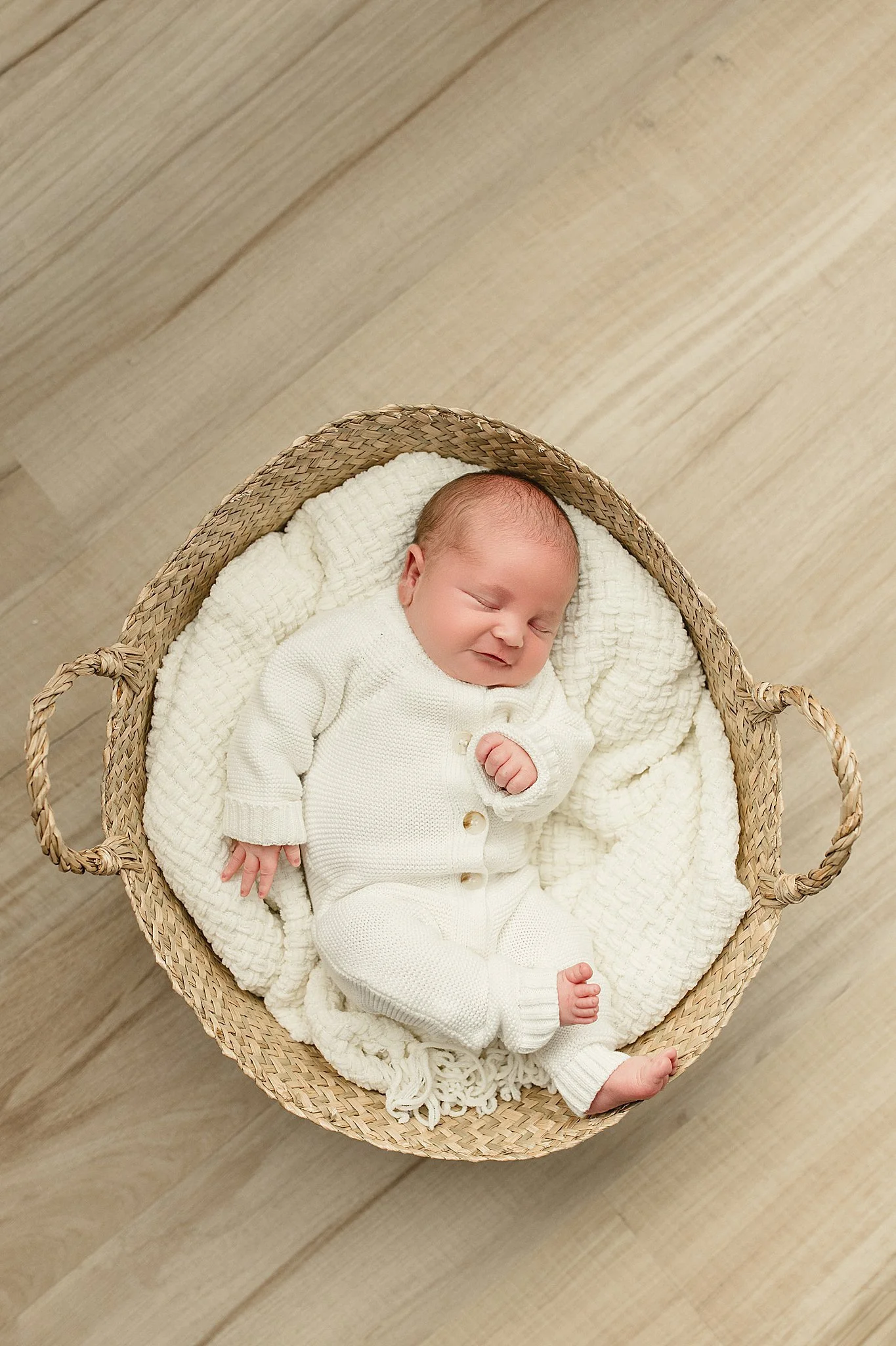 A newborn baby is sleeping in a wicker basket on a wooden floor, dressed in a white knitted outfit, with a cream-colored blanket inside the basket for a lifestyle newborn session near me.