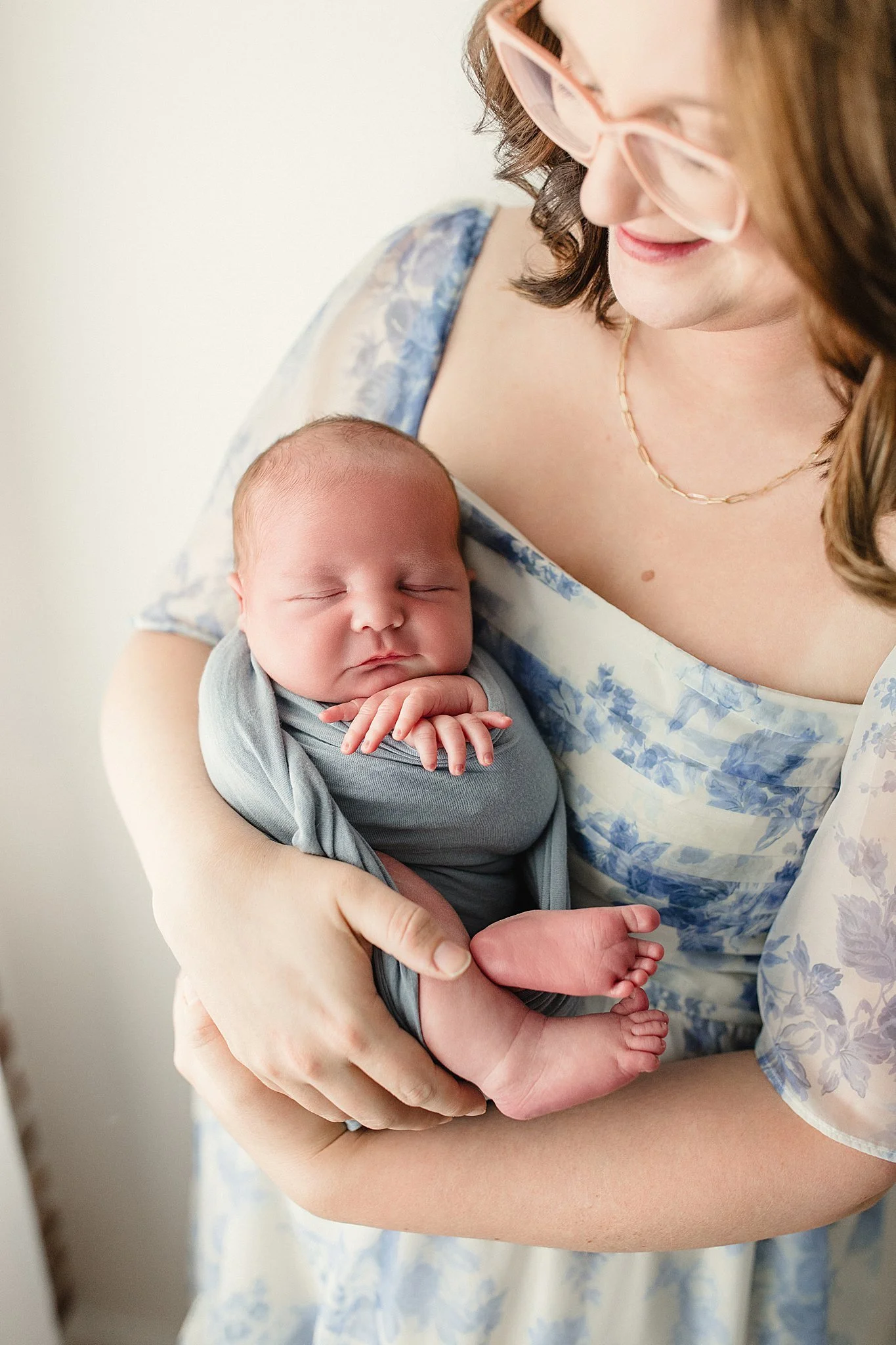 A woman holding a sleeping newborn baby against her chest, with the woman smiling gently and wearing pink glasses and a floral dress during a lifestyle newborn session in Naperville, Illinois.