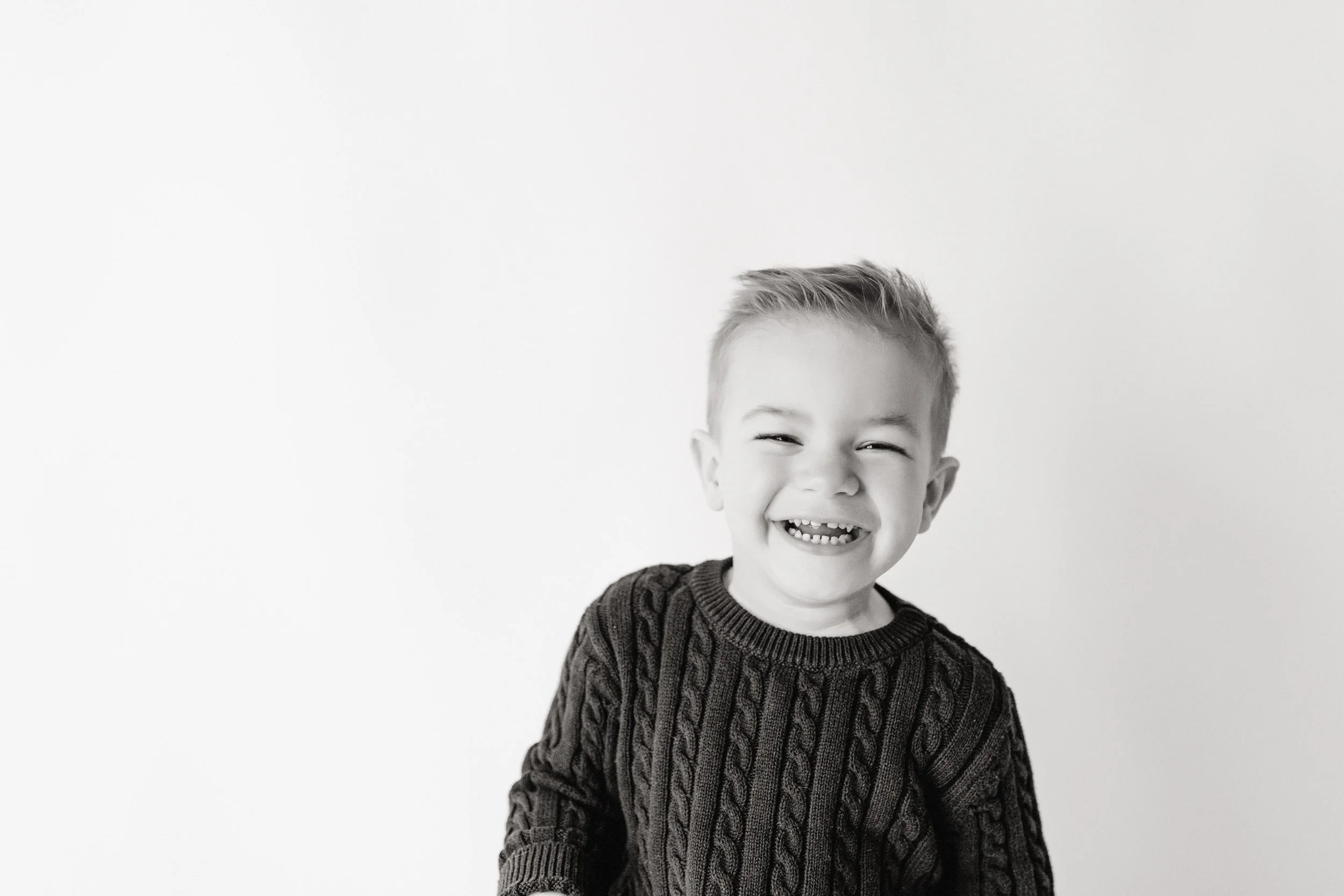 A smiling young boy with short hair, wearing a cable-knit sweater, standing against a plain white background during a studio photo session with Ally and B Photography in downtown Naperville, IL