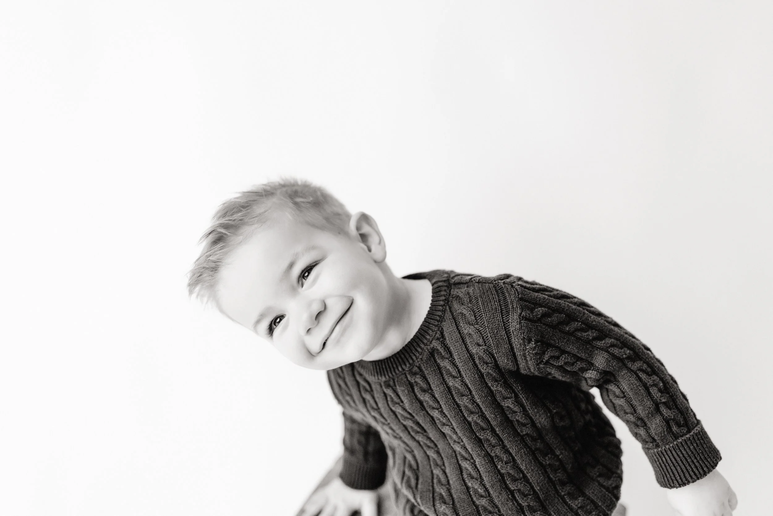 A smiling young boy with light hair wearing a dark, cable-knit sweater, leaning forward and captured in black and white against a plain background during a Naperville personality mini session with Ally and B Photography.