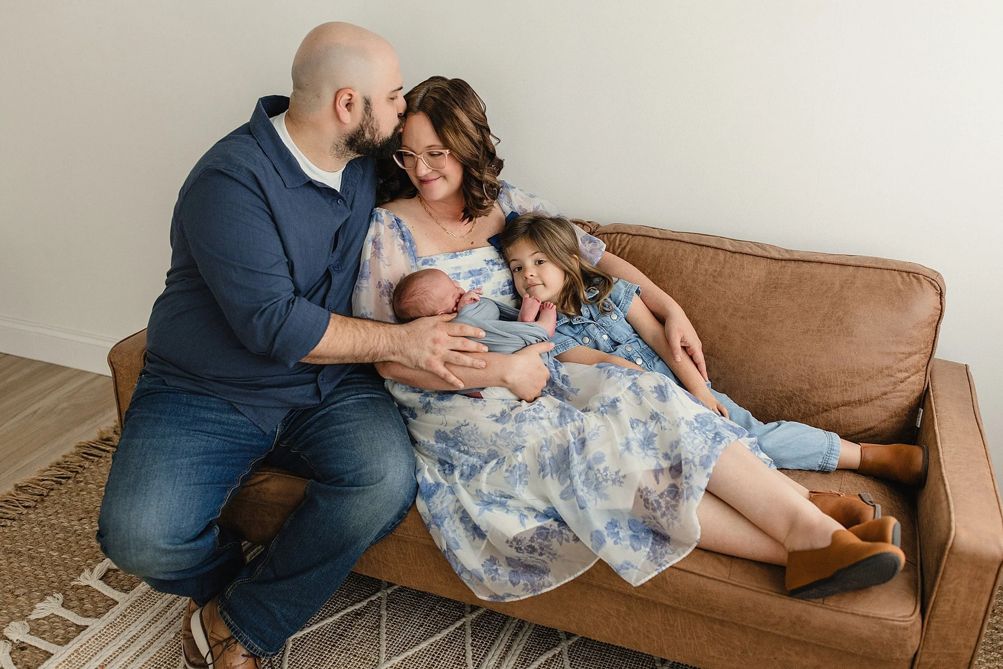 Family of four, including a man, woman, young girl, and newborn, sitting on a brown couch in a cozy room, sharing a tender moment in Naperville for a studio lifestyle newborn session.