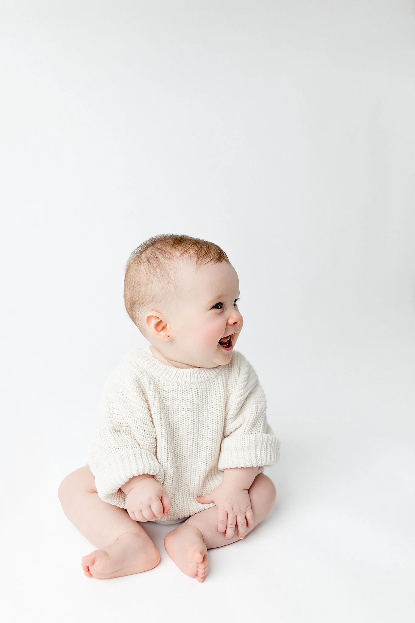 A happy baby sitting on a white surface, wearing a cream knit sweater, with a joyful expression during a simplistic baby milestone photography session with Ally and B Photography in Naperville.