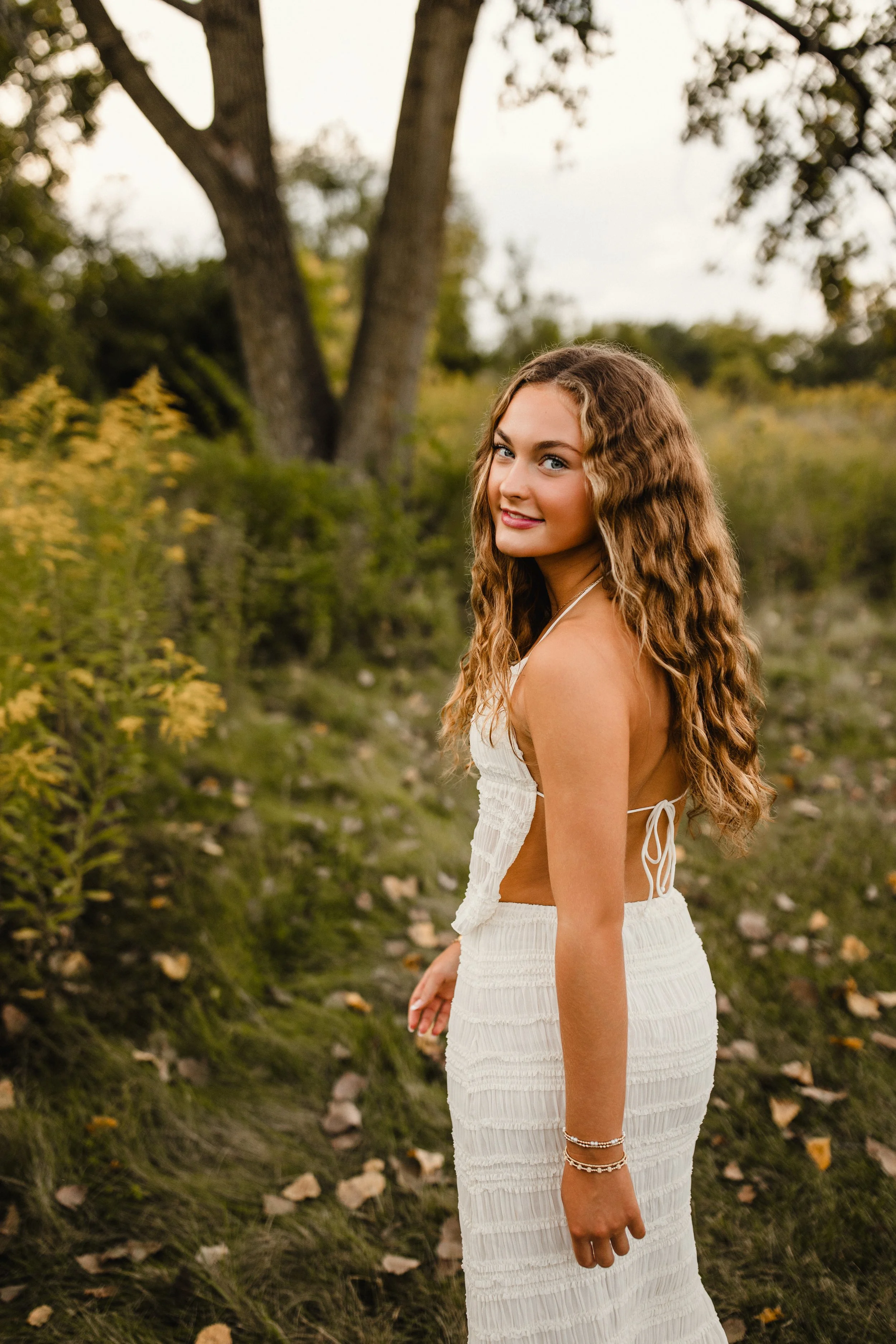 Young woman with wavy hair in a white dress standing outdoors in a natural setting with trees and bushes during her senior portrait session in Naperville for Naperville senior photography.