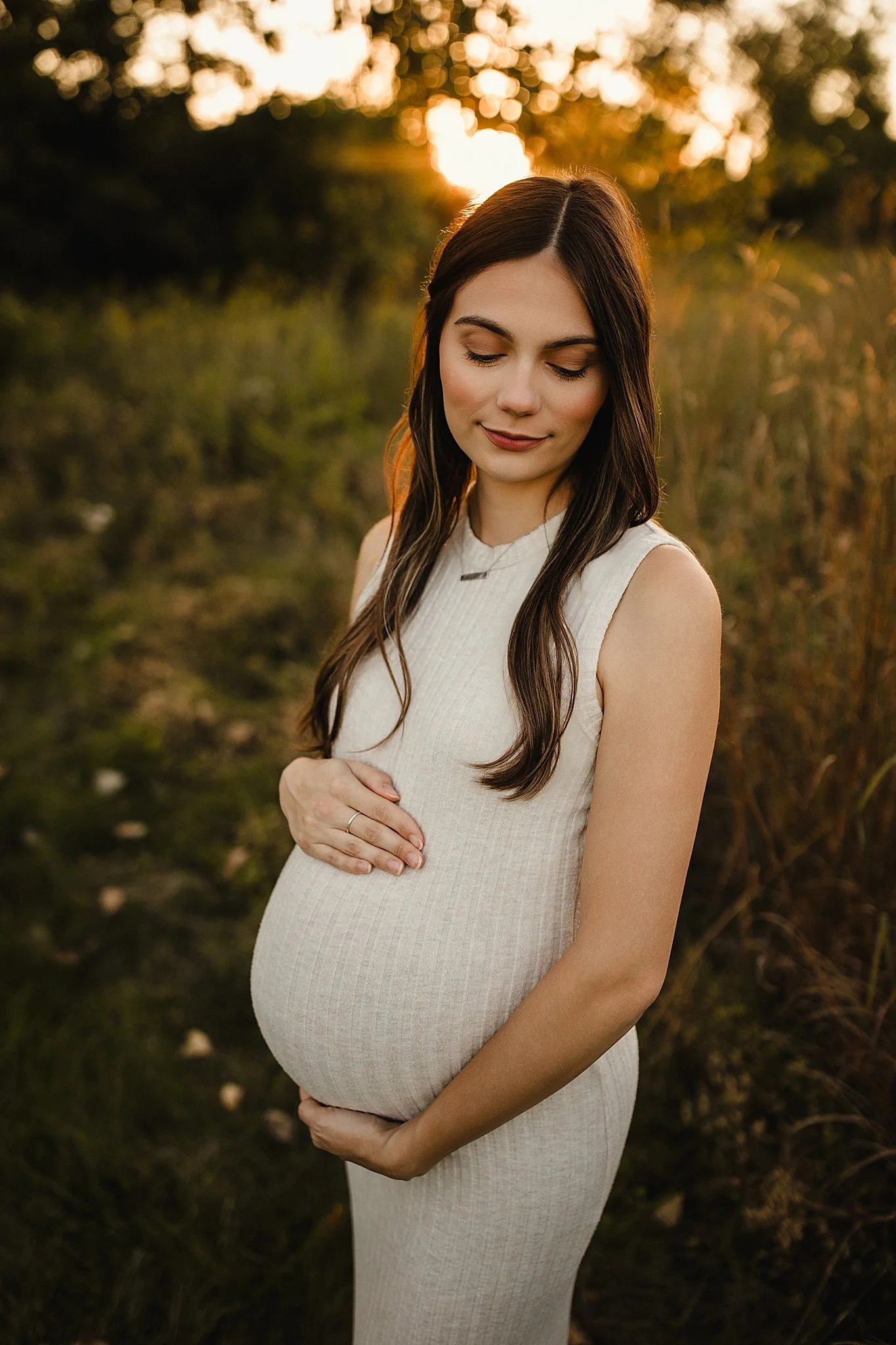 A pregnant woman standing outdoors during sunset, with her eyes closed and both hands resting on her belly, wearing a cream-colored sleeveless dress for Naperville maternity photos near Naperville.