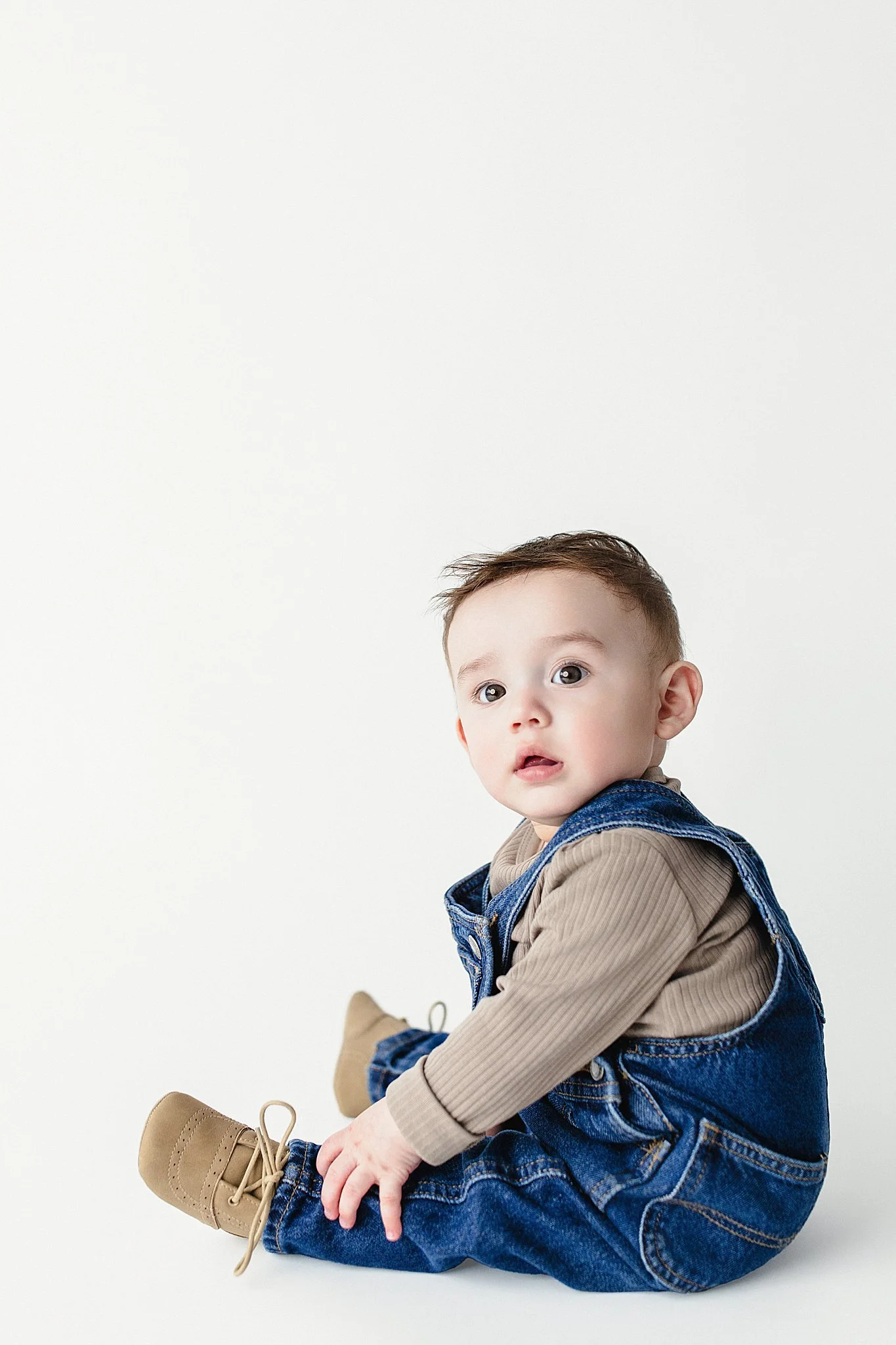 A young child with brown hair, wearing a beige long-sleeve shirt, denim overalls, and tan boots, sitting against a plain white background and looking at the camera with best baby photographer Naperville, IL.