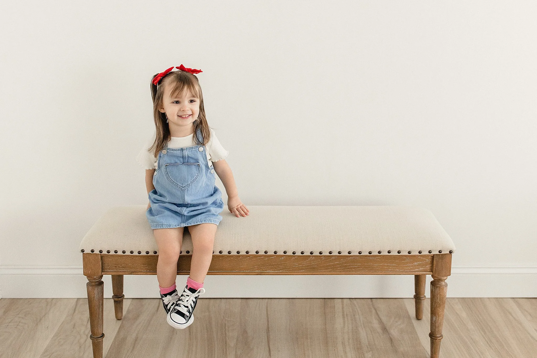 A young girl with a red bow in her hair sitting on a beige upholstered bench against a plain white wall. She is wearing a white shirt, denim overalls, pink socks, and black and white sneakers during a kids photoshoot near me.