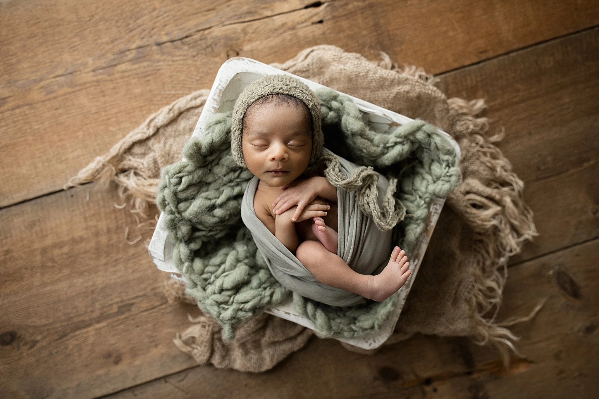 A newborn baby with dark skin and closed eyes, wrapped in a gray cloth, wearing a knit hat, and lying on soft blanket inside a small white box on a wooden floor with Naperville newborn photographers.