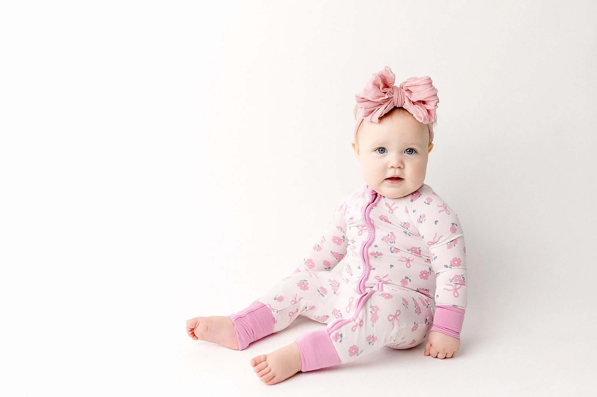 A baby girl with a pink headband and floral pajamas sitting on a white background for one year photos near me.