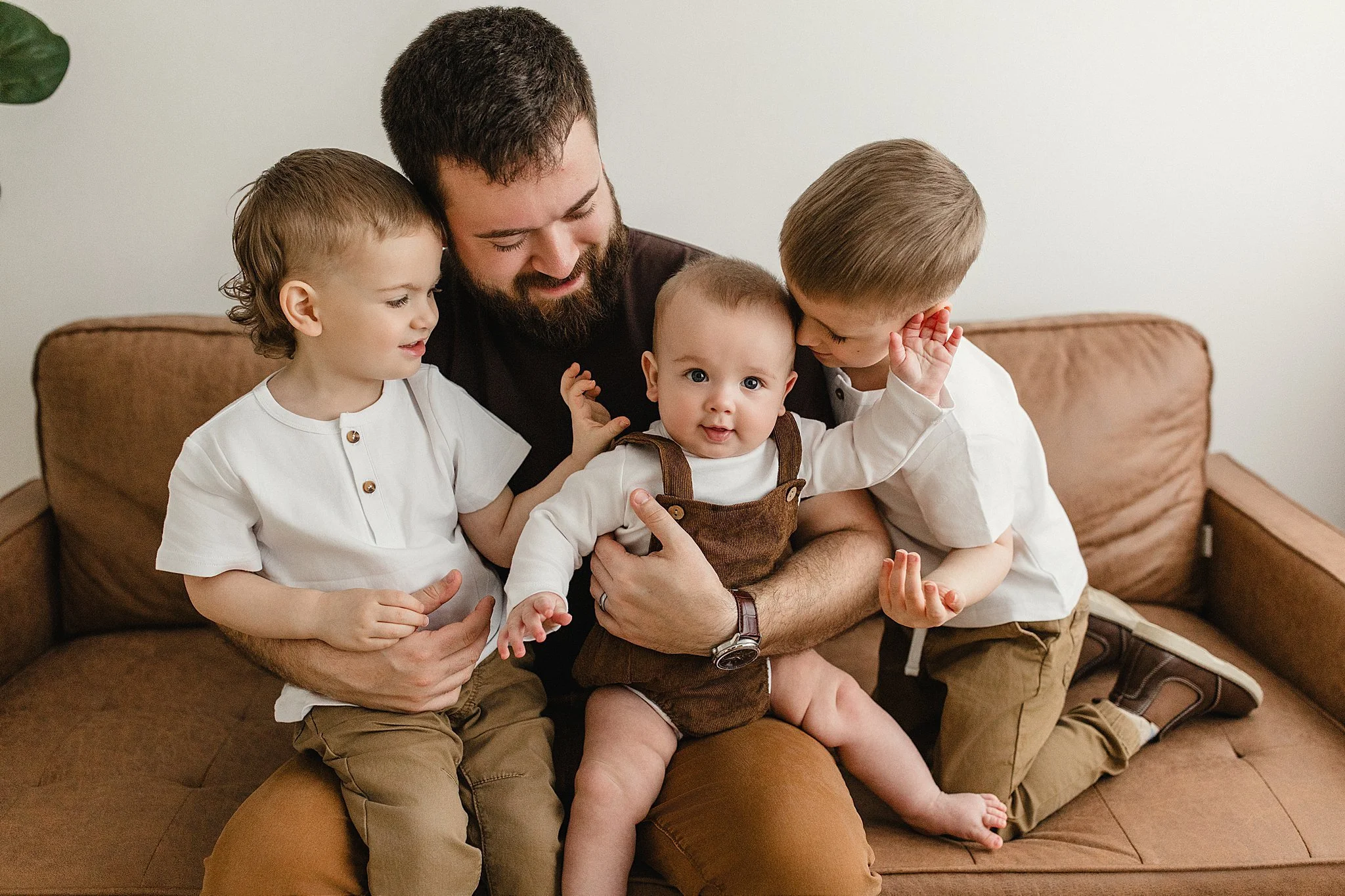 A man with three young children sitting on a brown couch, playing and smiling inside, with light-colored walls in the background during family photos near me.