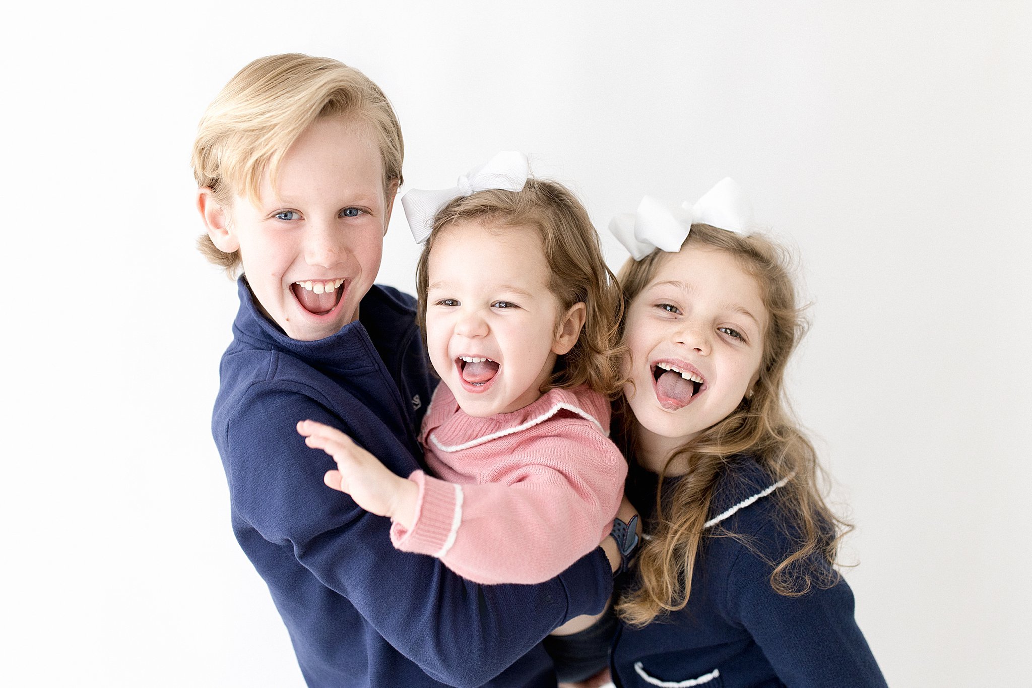 A happy boy holding two young happy girls, all with light-colored hair and white bows, smiling and laughing against a white background with the best kids photographer in Naperville, IL.