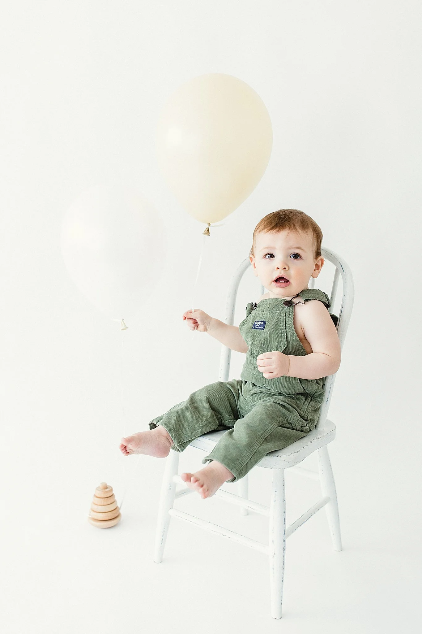 A young child sitting in a white chair holding a cream-colored balloon, with two more balloons floating nearby, against a plain light background in milestone session in Naperville, IL.
