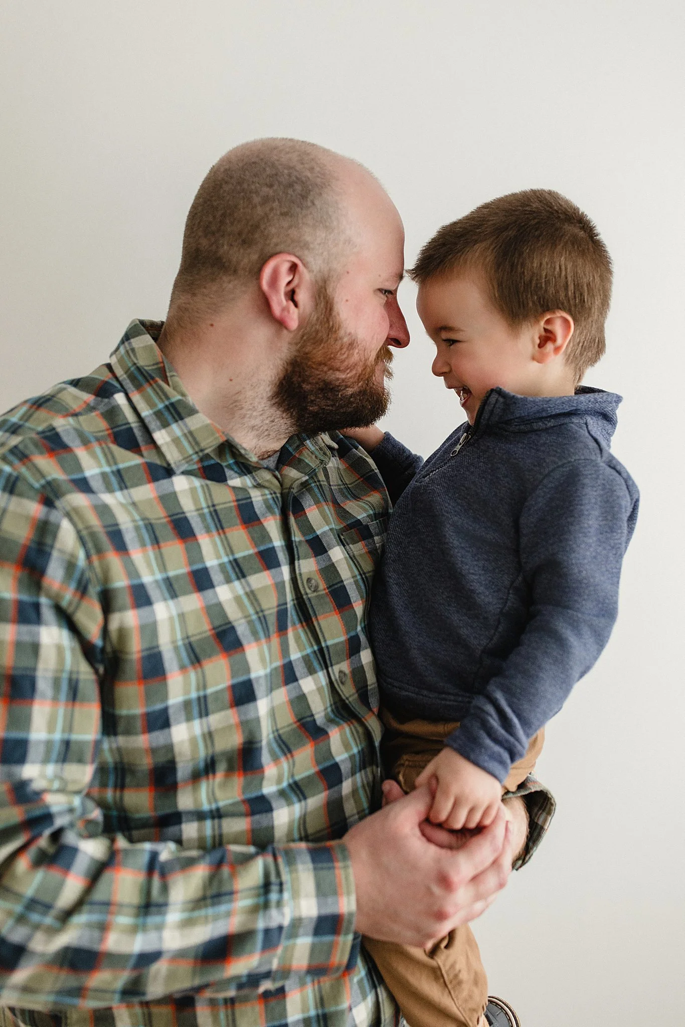 A man with a beard and a plaid shirt holding a young boy in a navy jacket, both smiling and touching foreheads in a close-up indoor shot during family photos with Ally and B Photography.
