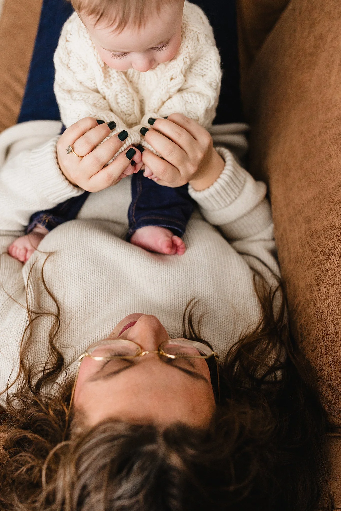 A woman with curly hair and glasses lying down on a couch, smiling, while a small child sits on her stomach, holding her hands and looking down at her for an in studio lifestyle family photo session in downtown Naperville, IL.