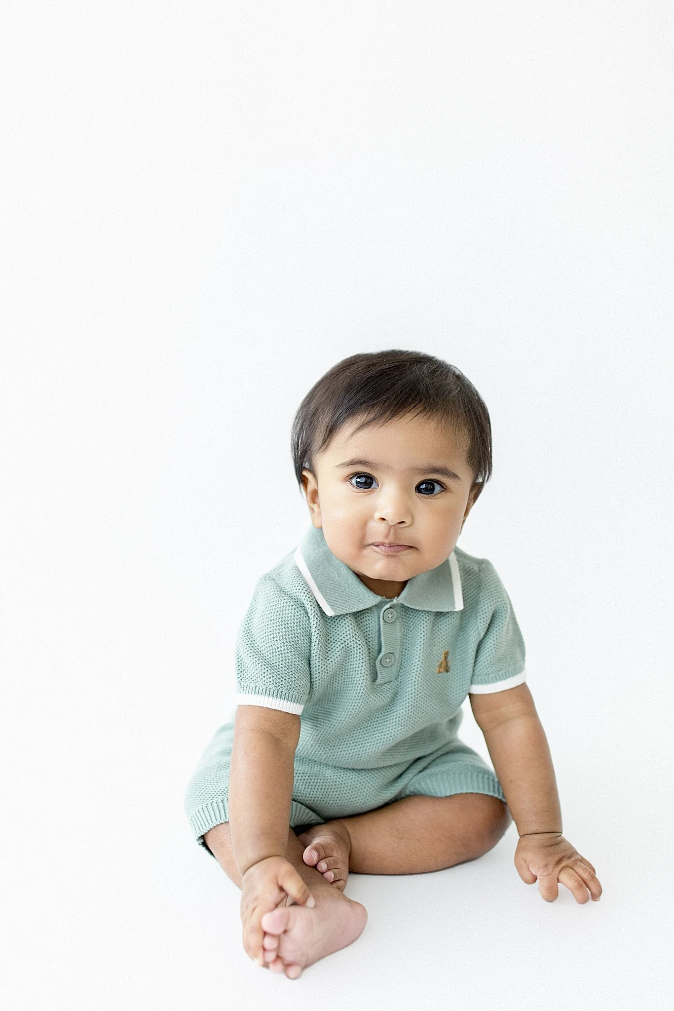 A young child with dark hair and big eyes sitting on a white background, wearing a light green polo shirt and matching shorts with the best baby photographer in Naperville, IL.