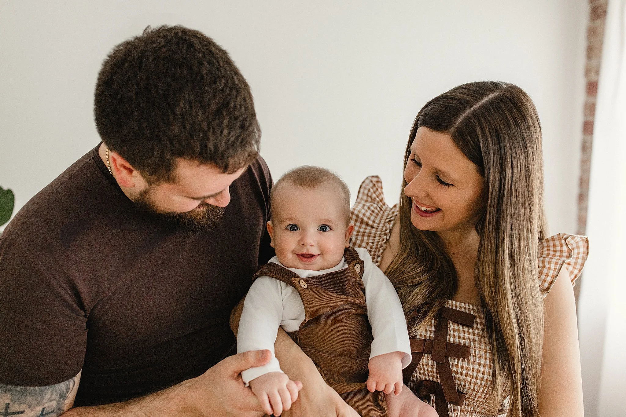 A family of three, a man, a woman, and a baby, sitting together indoors. The baby is smiling and being held by the man, while the woman looks at the baby with a smile. The scene is warm and cheerful during a family photo session in Naperville, IL.