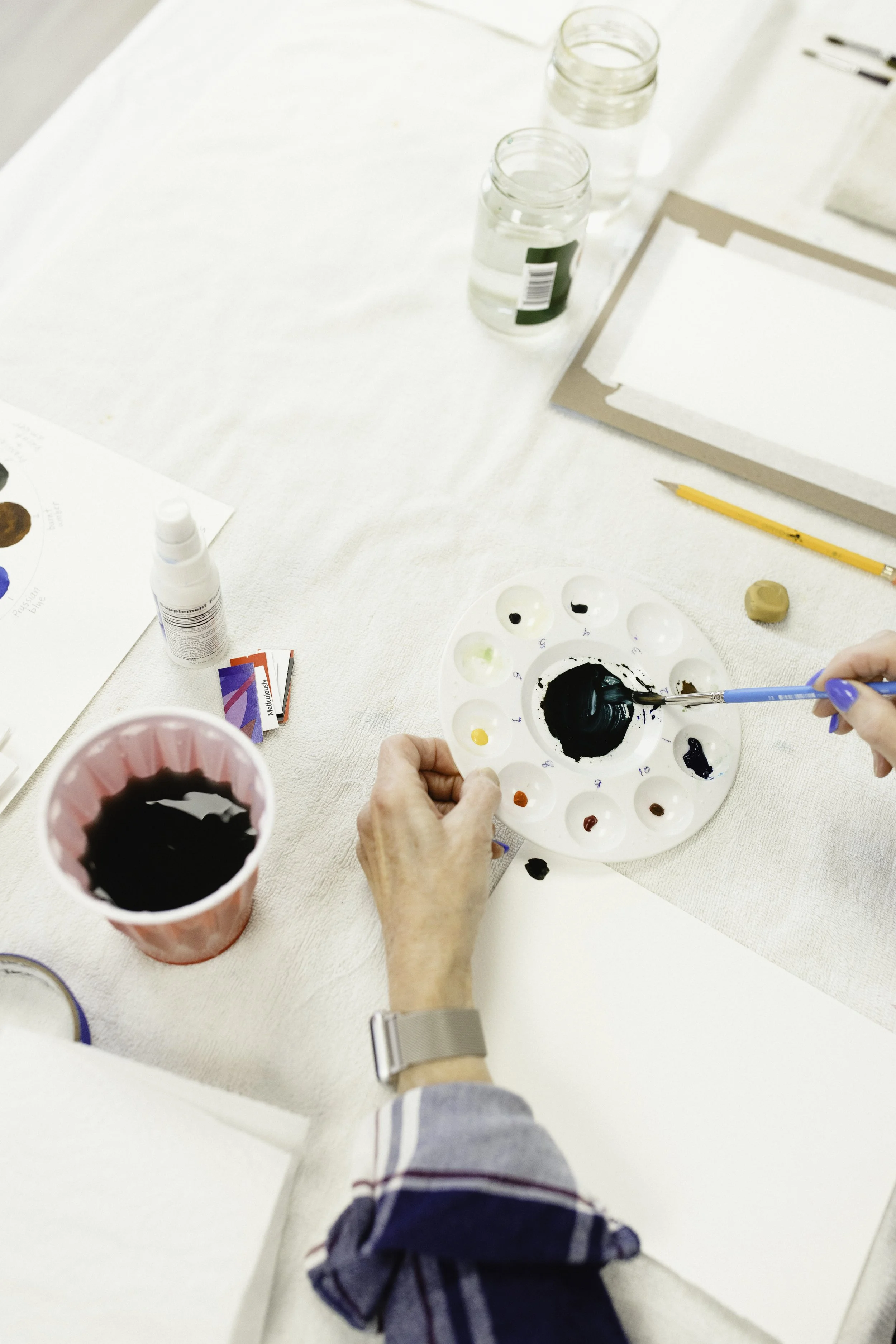 Person mixing black paint on a circular palette surrounded by art supplies on a white table during beginner watercolor class in Naperville, IL, taught by Jonnette Maslowski.