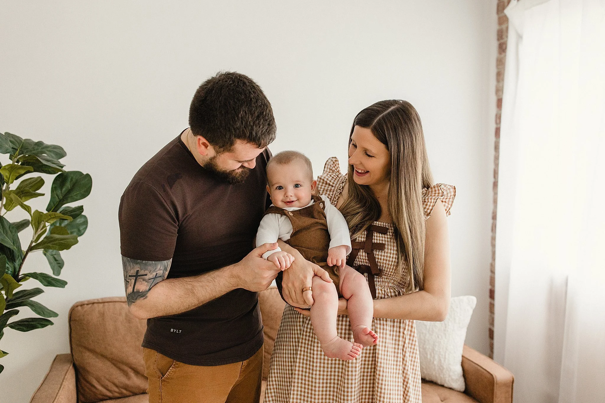 A happy family of three, including a man, woman, and baby, indoors in a living room with a beige couch and large window. The man holds the baby, and the woman smiles at the baby during family photos in Naperville, IL.
