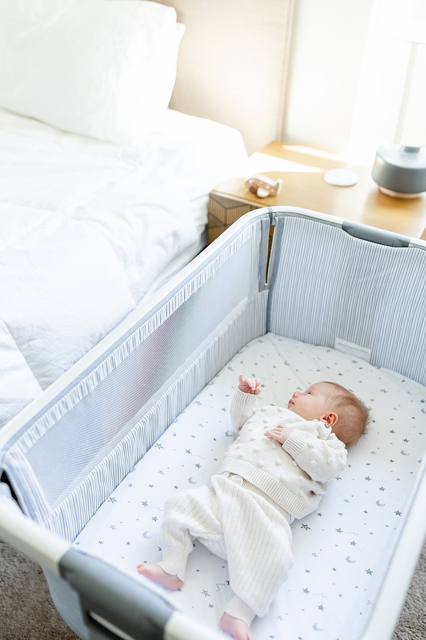 A baby lying in a crib with star and moon patterned sheets, wearing a cream-colored sweater and pants, reaching up with one hand, with a changing table and a lamp on a nightstand in the background during lifestyle newborn photos Naperville, IL.