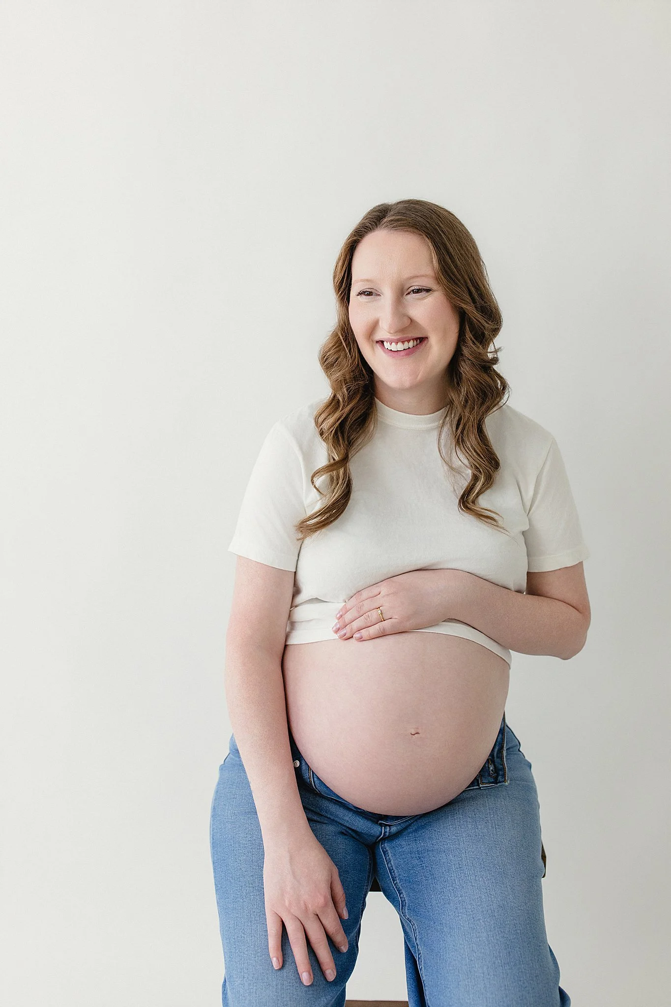 A pregnant woman with long wavy brown hair smiling and wearing a white t-shirt and blue jeans, holding her belly with her left hand, standing against a plain light-colored wall with the best maternity photographers near me.