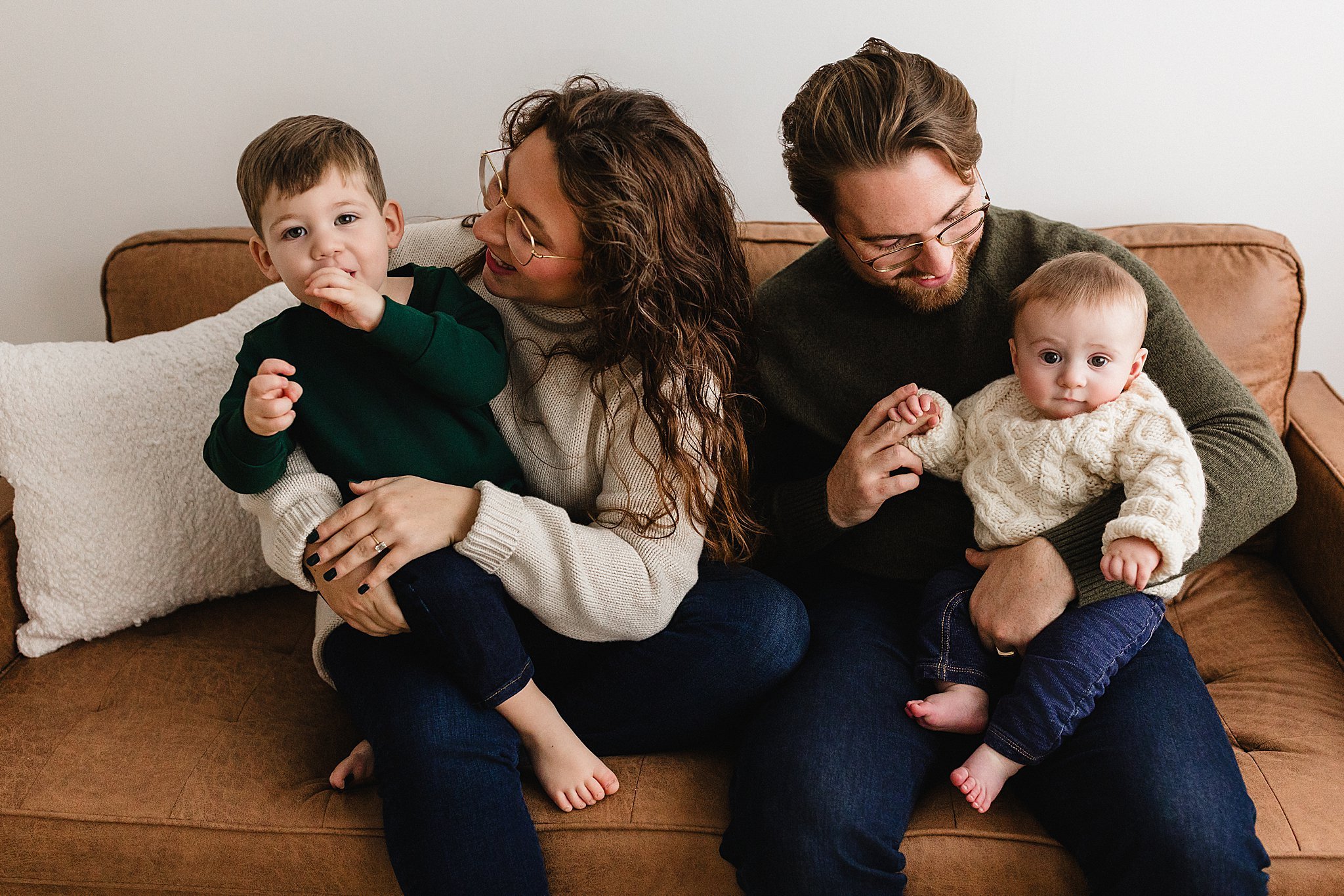 A family of four sitting on a brown couch, with a woman holding a young boy and a man holding a baby, all smiling and engaging each other for studio photos in downtown Naperville.