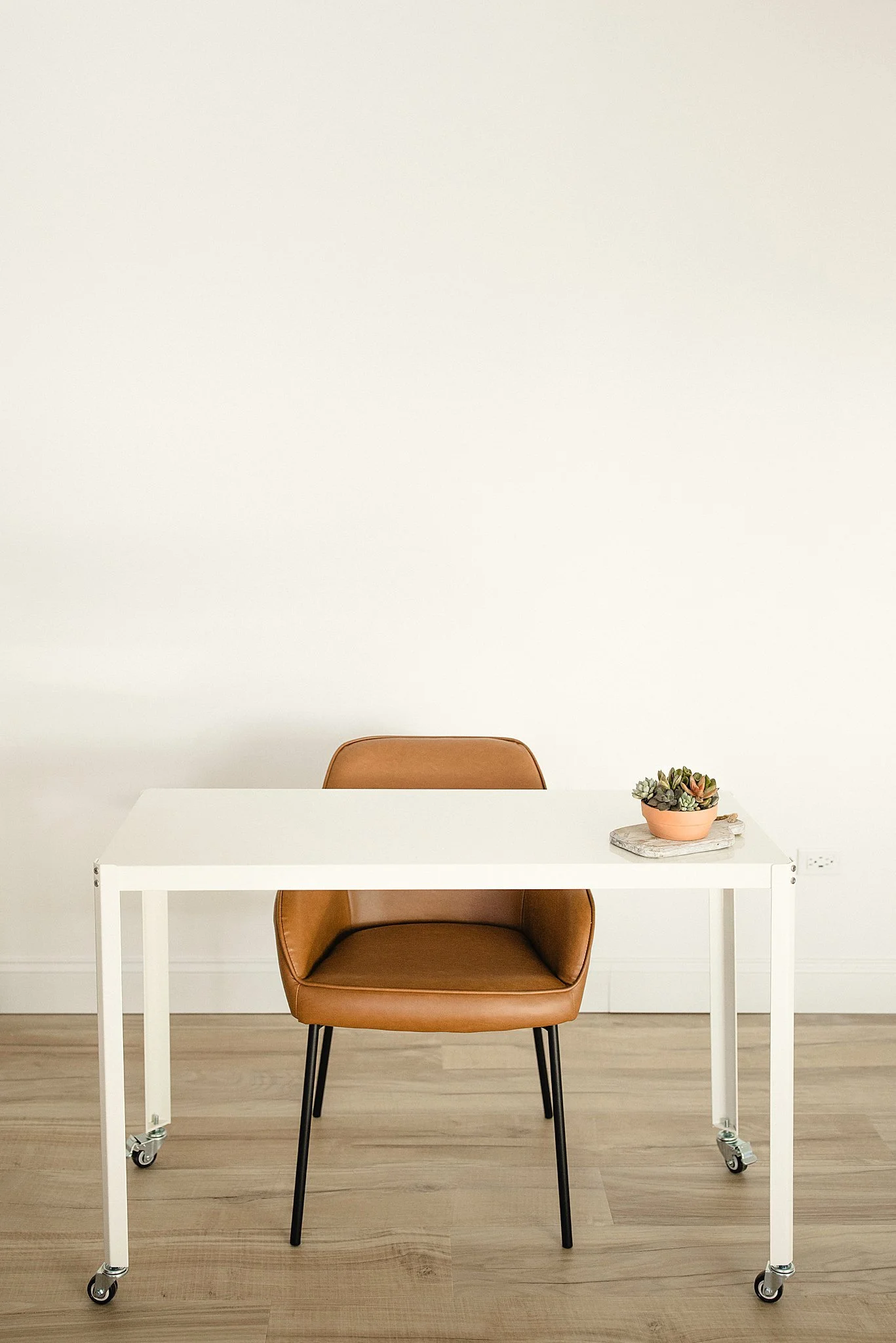 A white desk with casters is placed in front of a brown chair. On the desk, there is a terracotta pot with succulents on a white tray. The background is a plain white wall in photography studios near Naperville.