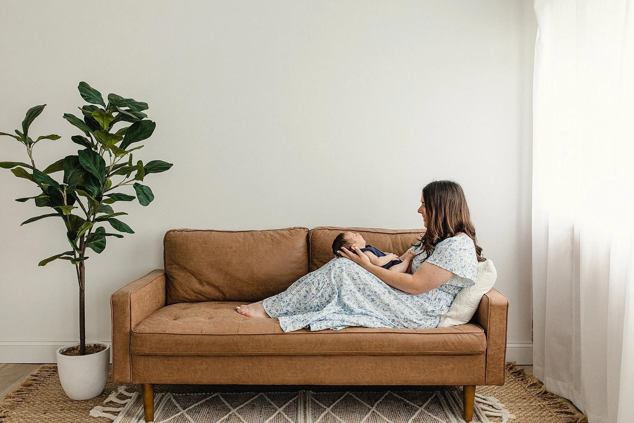 A woman with brown hair and a child lying on a tan couch in a room with white walls and a large green houseplant with lifestyle photographers Naperville.