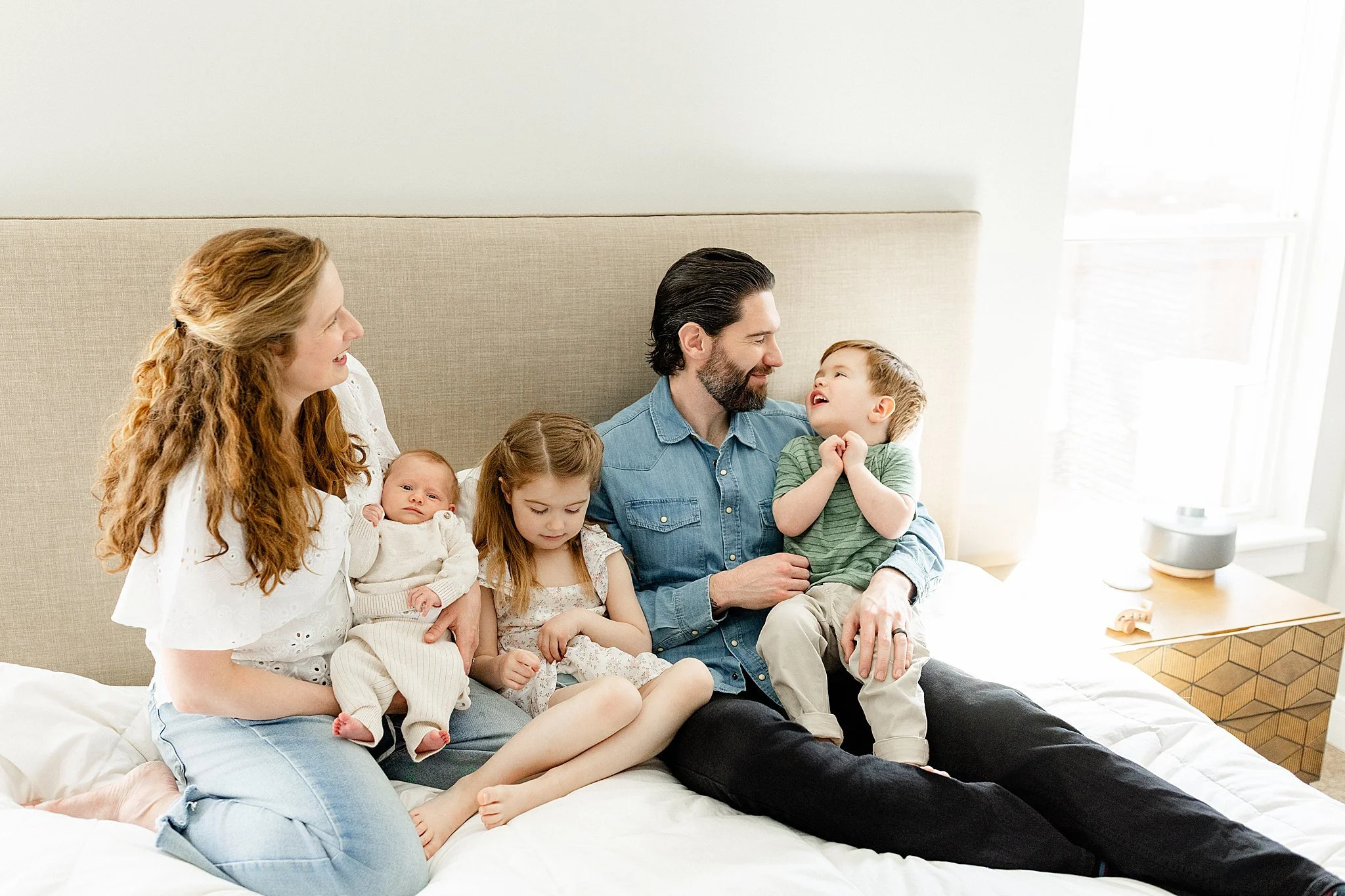 A family sitting on a bed, with two adults and four children, all smiling and enjoying each other's company in a bright, modern bedroom with a lifestyle newborn photographer near me.