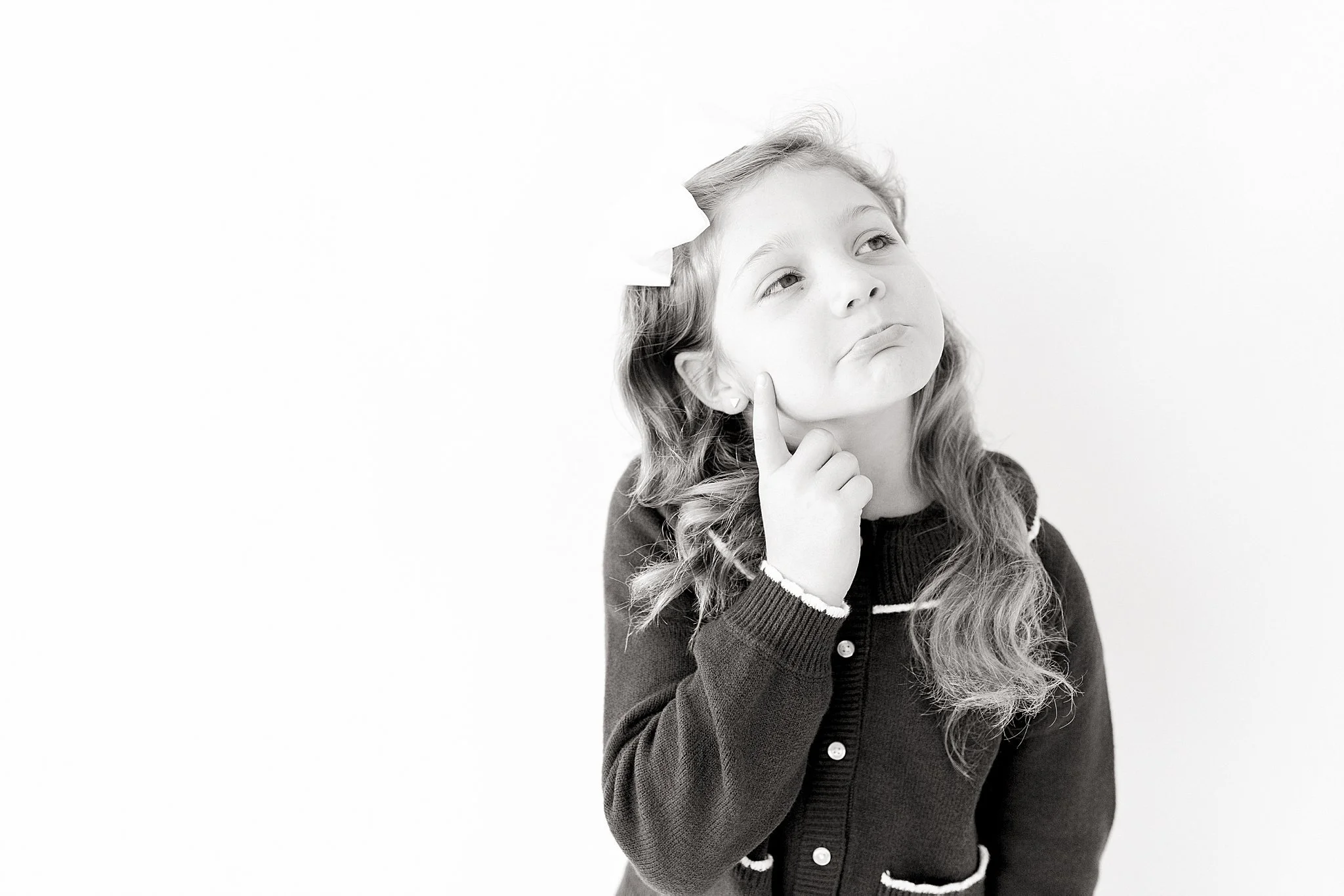 A young girl with long wavy hair, wearing a dark sweater, is touching her cheek with her finger and looking upwards with an inquisitive expression during personality portraits near Chicago, IL.