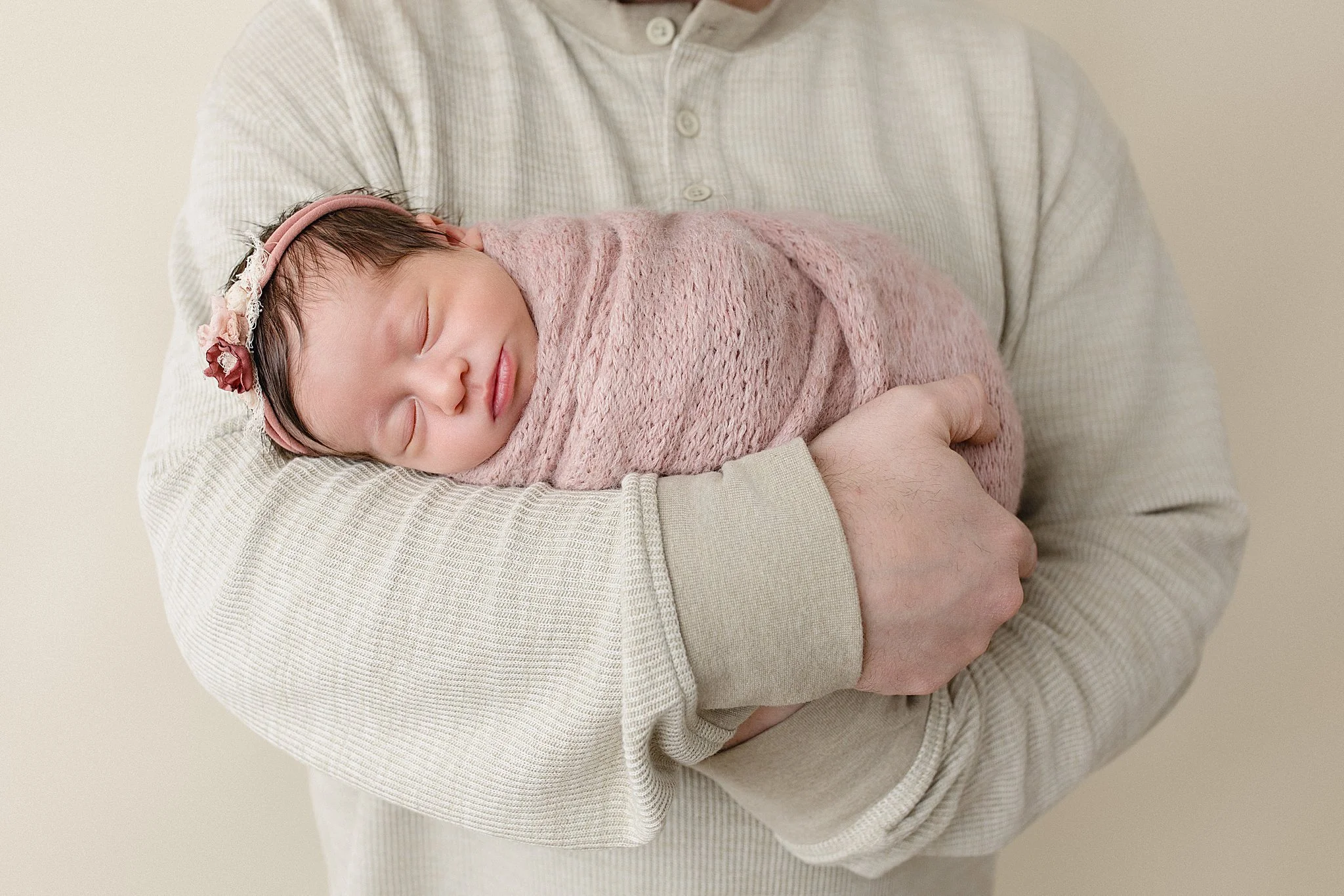 A baby girl wrapped in a pink blanket with a pink headband, peacefully sleeping in an adult's arms during newborn photos with Naperville newborn photographer.