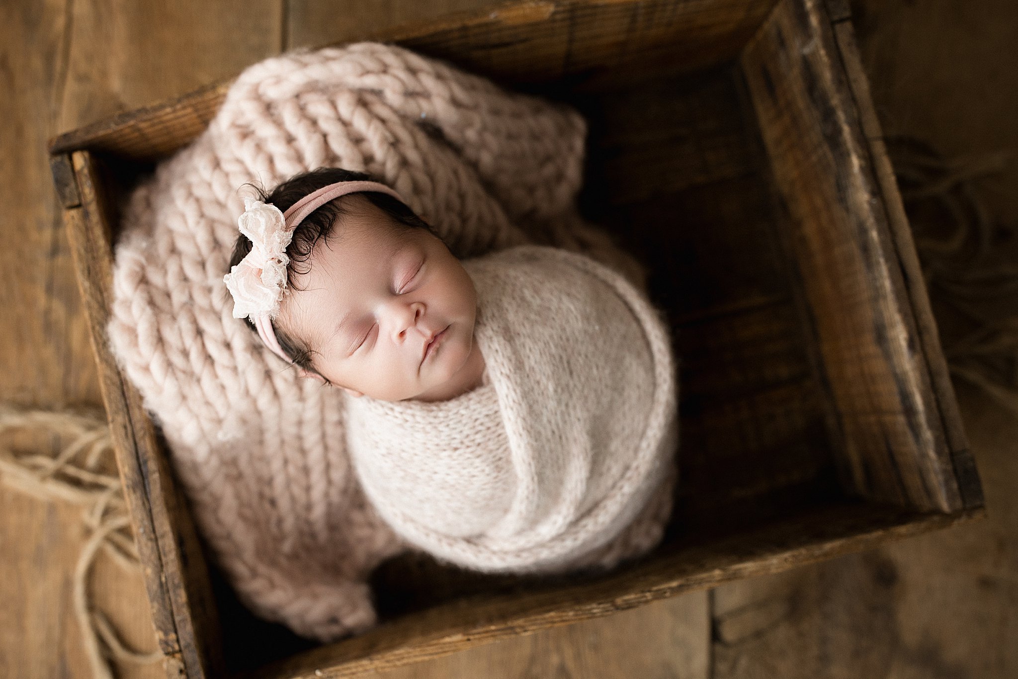 A sleeping baby with dark hair, wearing a pink headband with a bow, wrapped in a cream knit blanket, lying in a wooden box with a chunky cream knit pillow during posed newborn photos Naperville.