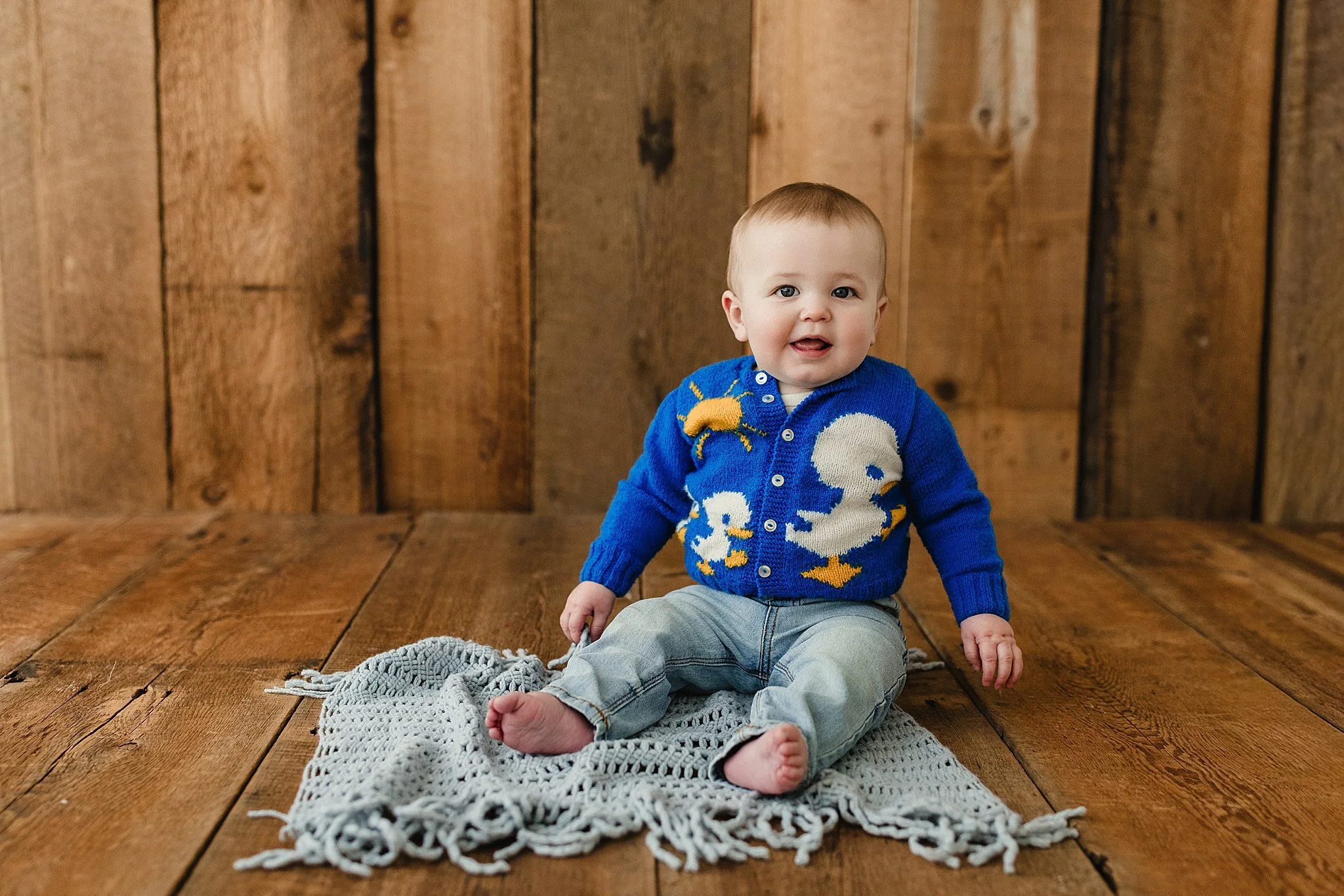 A smiling baby sitting on a gray knitted blanket on a wooden floor, with a wooden wall background, wearing a blue sweater with duck and swan designs and light blue jeans during milestone photos with Ally and B Photography in downtown Naperville, IL.