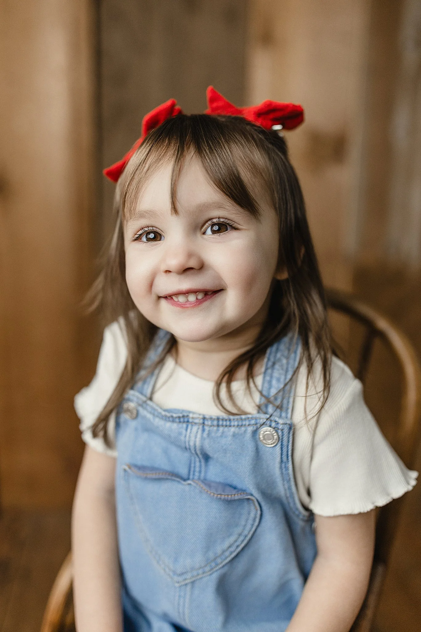 A young girl with brown hair, light skin, and a red bow on her head, smiling while sitting on a wooden chair. She is wearing a cream-colored t-shirt and denim overalls for a kids milestone photoshoot.