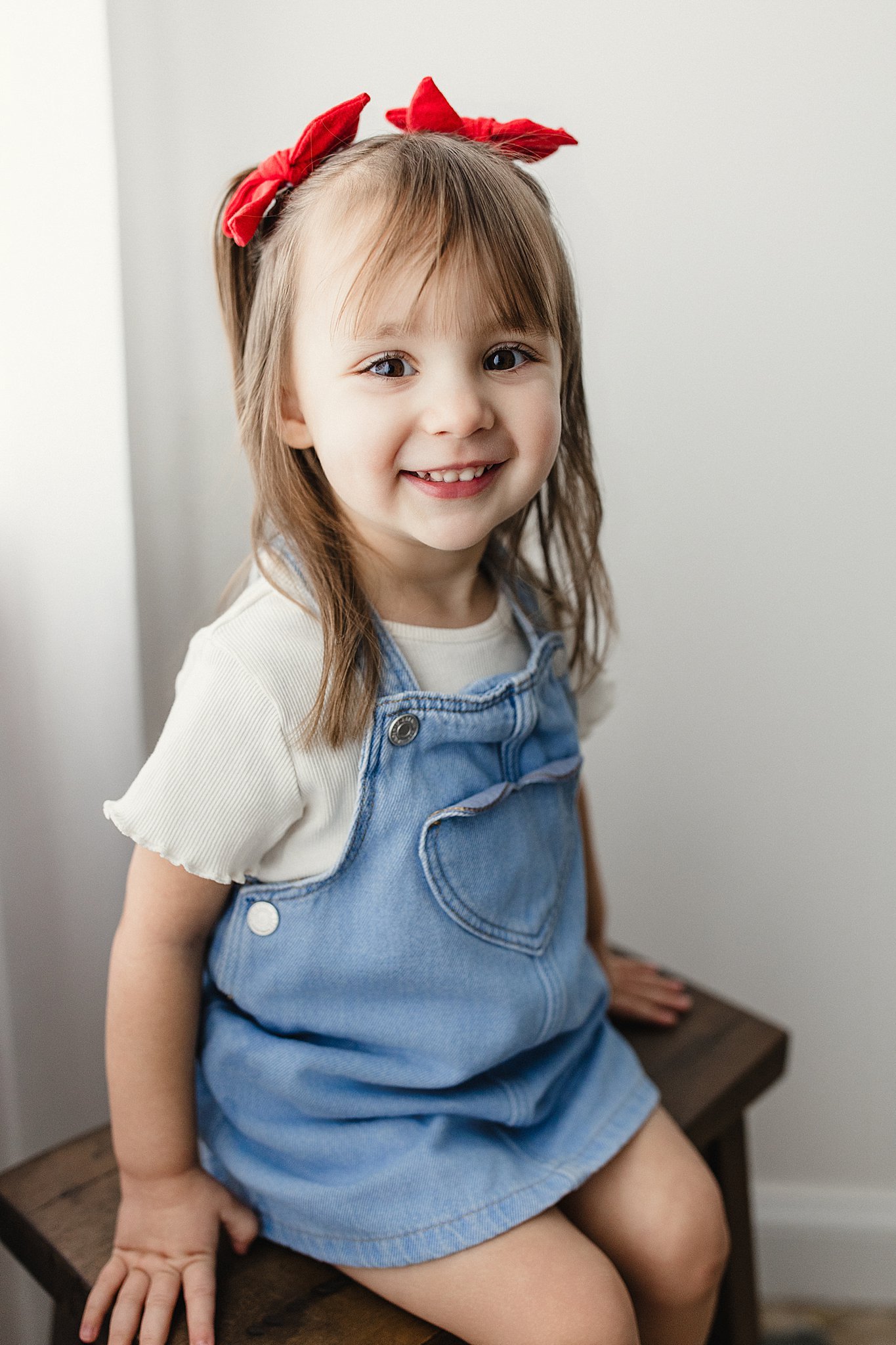 A young girl with brown hair, wearing a red bow, a cream-colored shirt, and denim overalls, sitting on a wooden stool and smiling at the camera during a kids photoshoot in Naperville, IL.